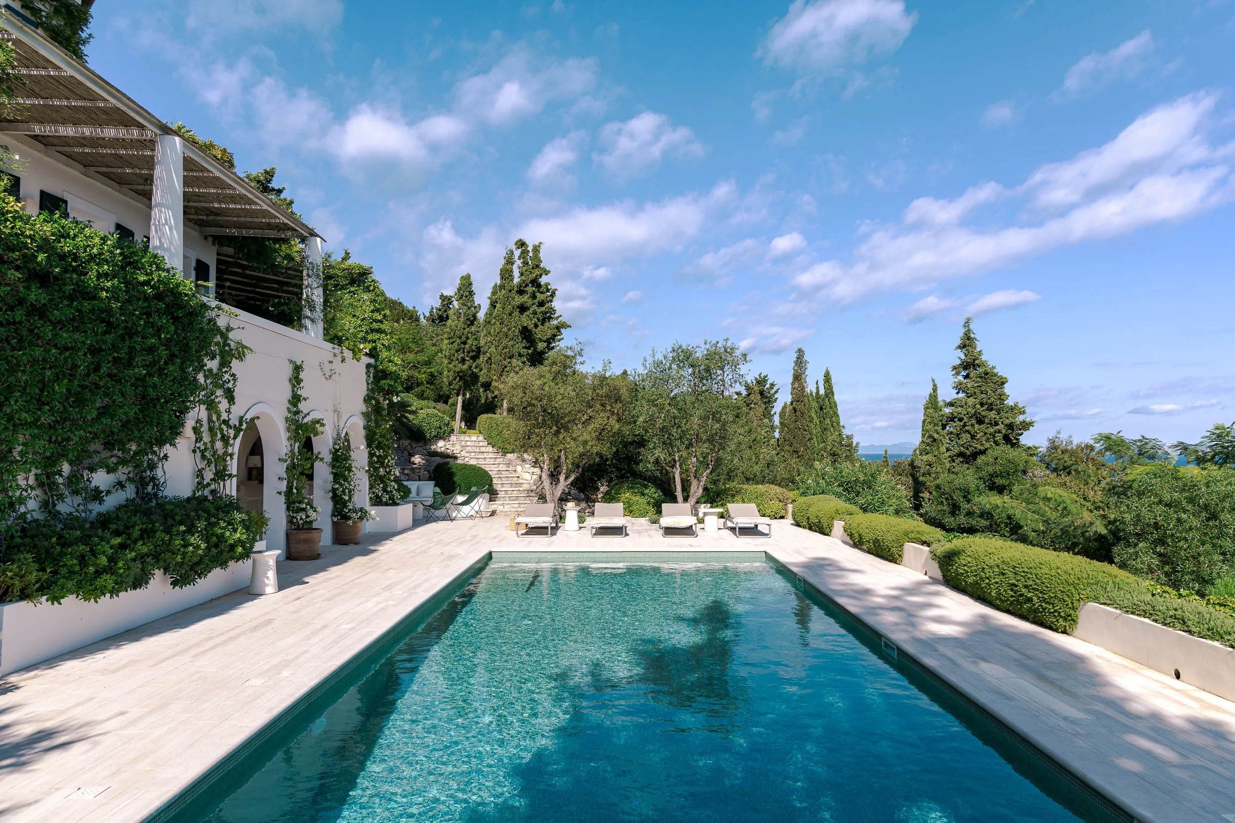Luxury swimming pool area with lounge chairs, surrounded by lush greenery and trees, under a bright blue sky with scattered clouds.