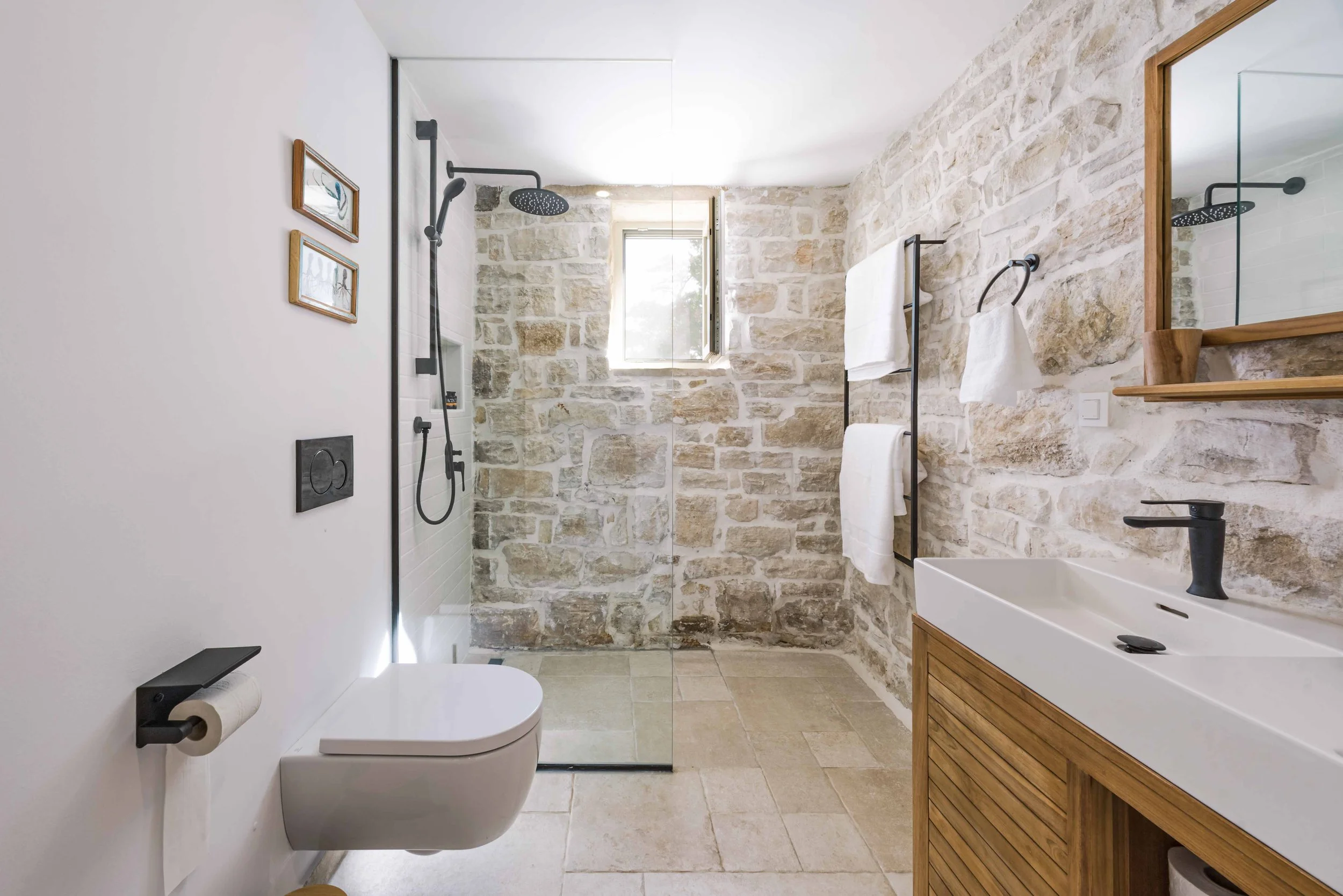 Modern bathroom with stone walls, walk-in shower with glass partition, white toilet, black fixtures, and wooden accents