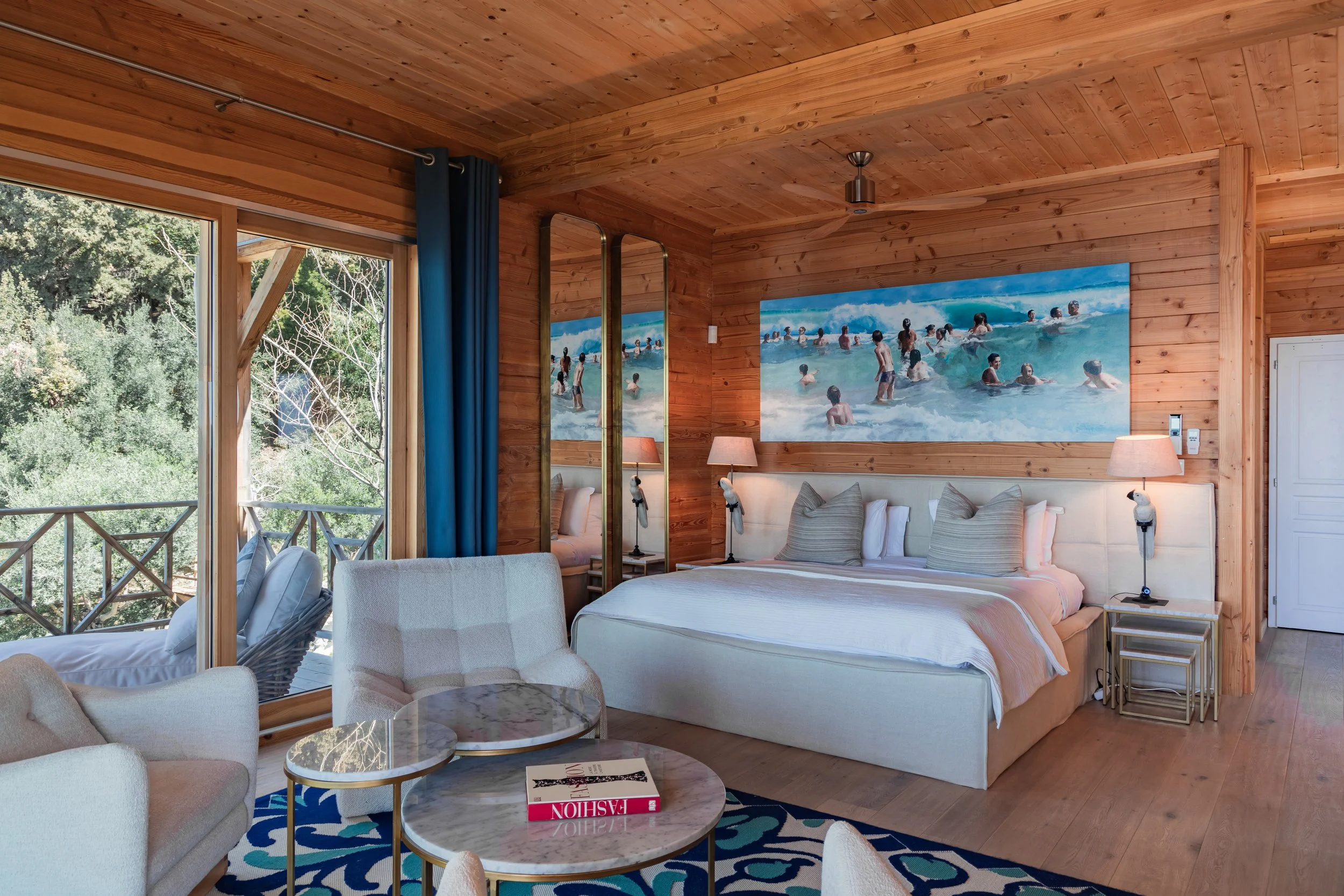 Hotel room with wooden walls and ceiling, large bed with pillows, side tables with lamps, a colorful patterned rug, a round marble coffee table with a book, a beige armchair, a sliding glass door leading to a balcony with greenery outside, and a larg