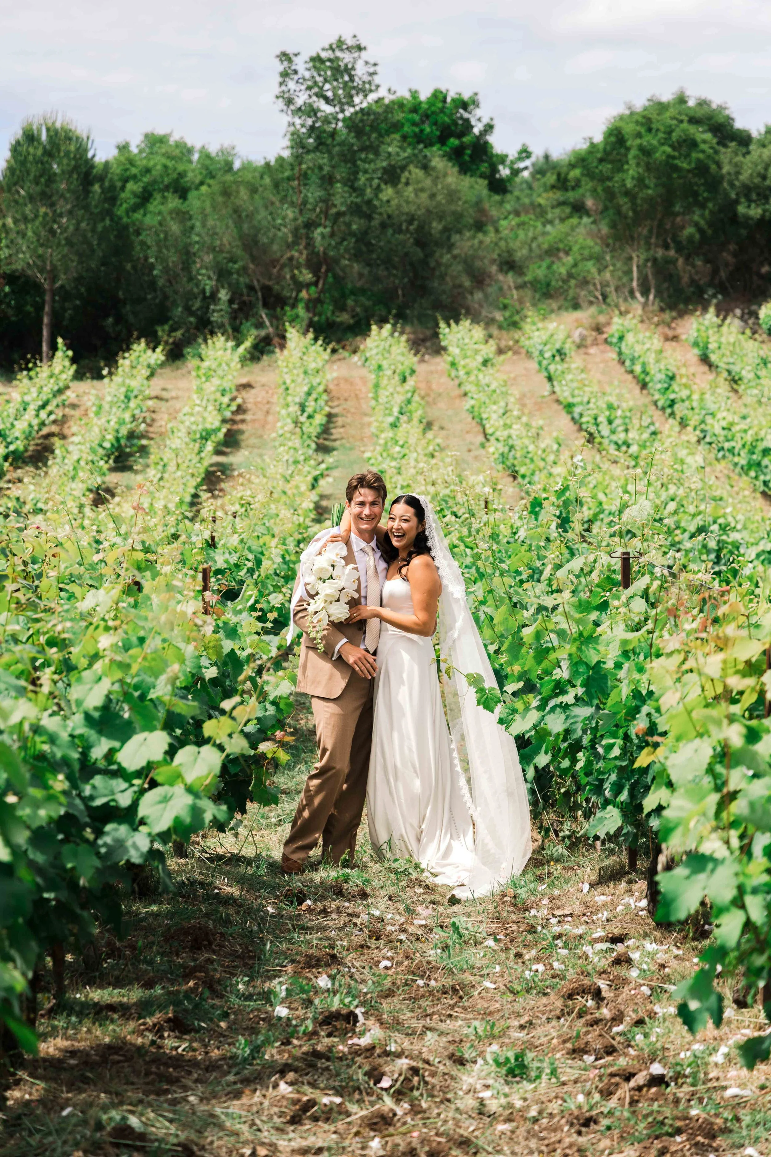 A newlywed couple smiling and laughing in a vineyard with lush green grapevines and trees in the background, the bride holding a bouquet and wearing a white wedding dress with a veil, the groom in a beige suit.