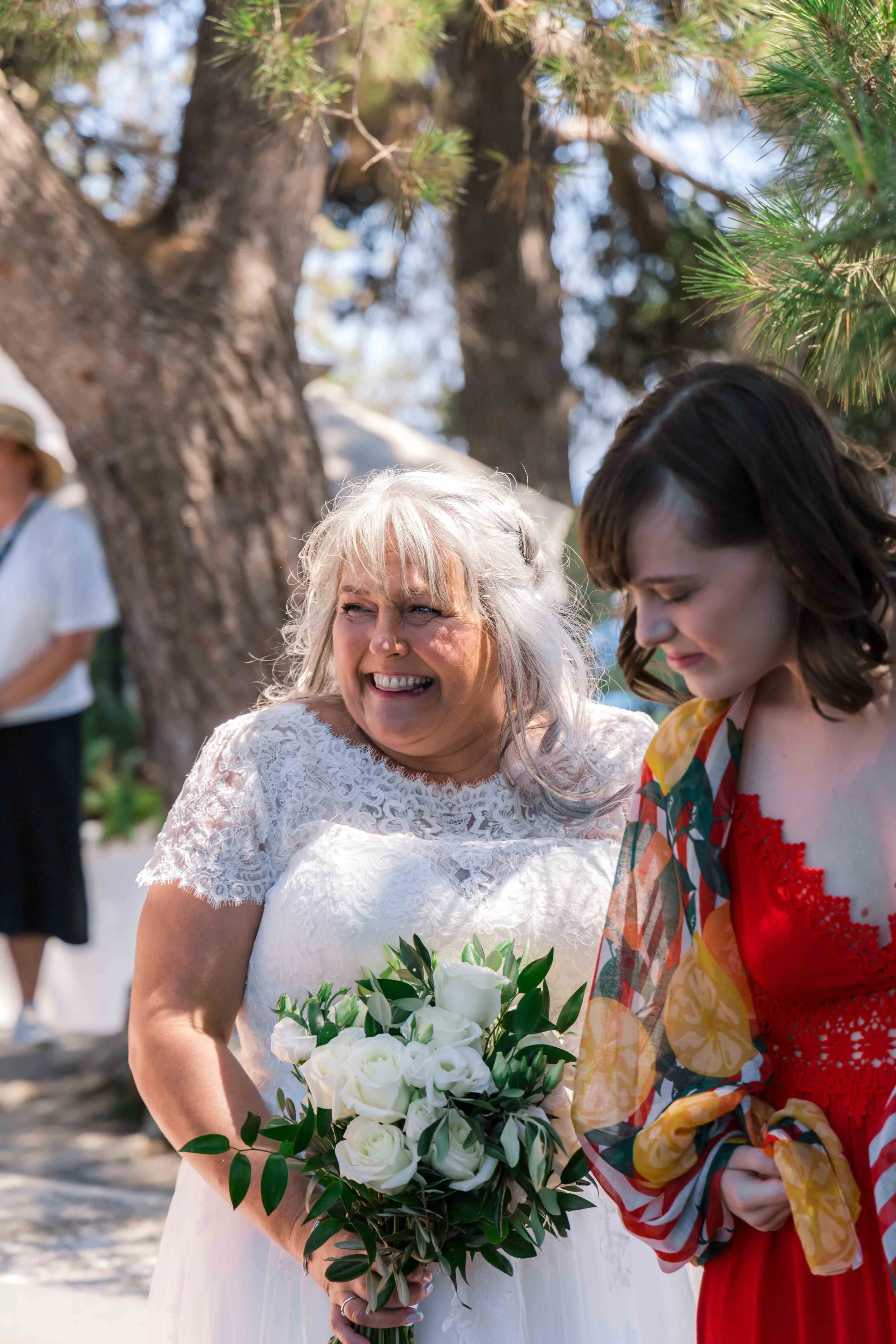 A woman with gray hair in a white lace dress holding a bouquet of white roses is smiling, standing outdoors near a tree, with another woman in a colorful dress beside her.