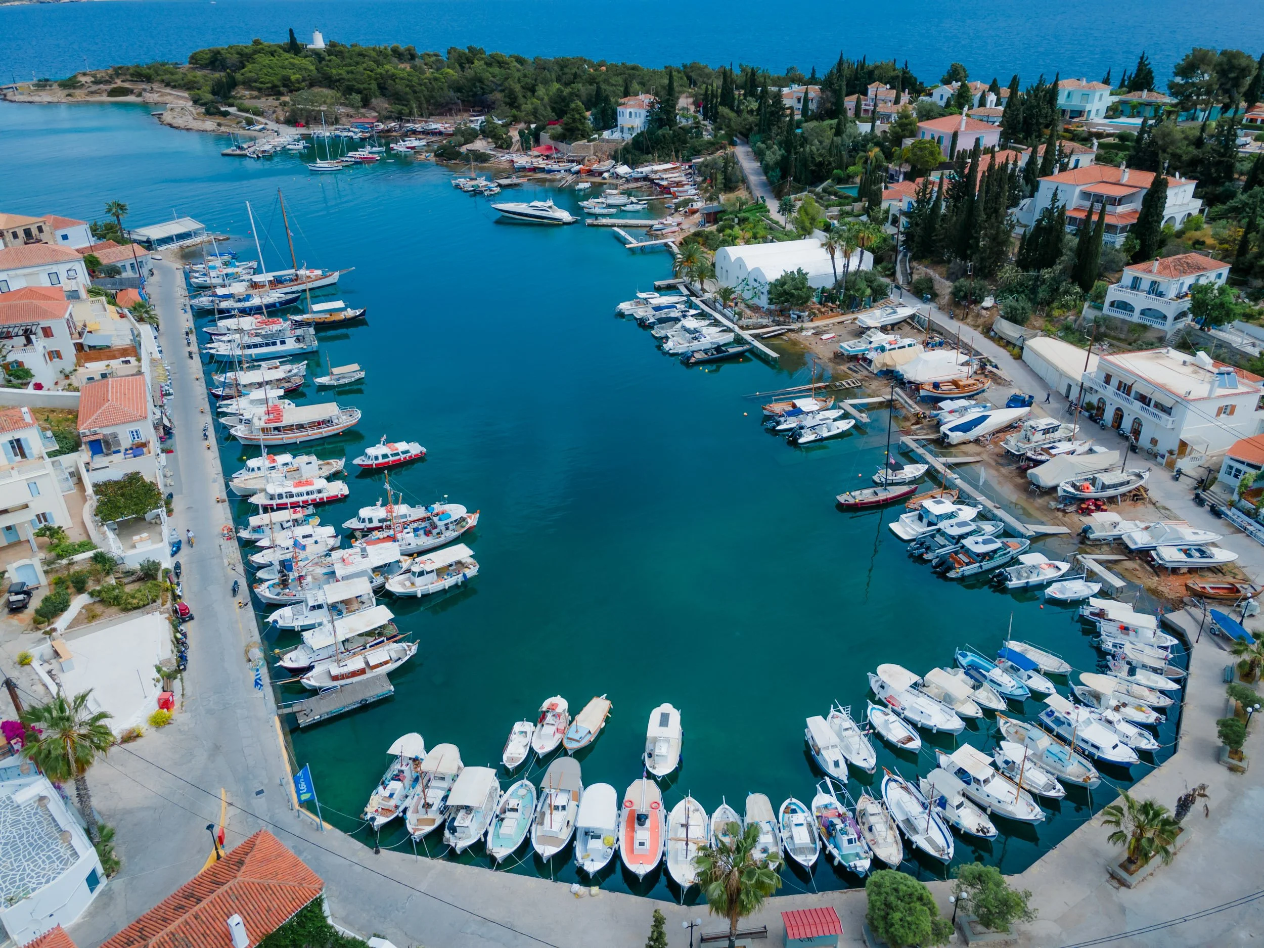 Aerial view of a marina with numerous boats docked along the sides, surrounded by white buildings with orange tiled roofs and green trees, with the sea in the background.