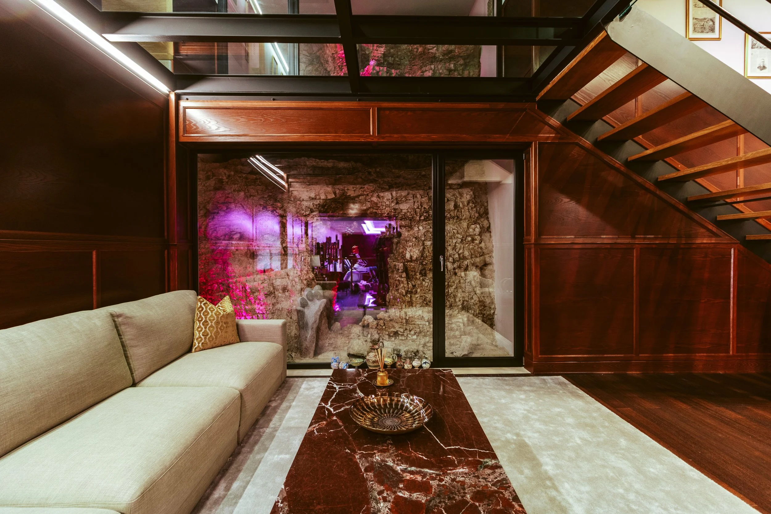 Living room with beige sofa, marble coffee table, wooden paneled walls, glass sliding door, staircase with wood steps and black risers, stone wall visible through the glass, framed pictures on the wall, plush rug on the floor.