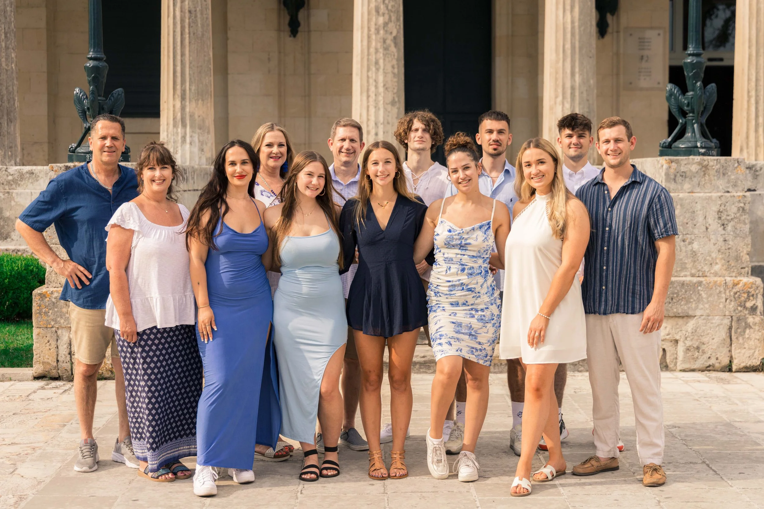 Group of 14 young adults and middle-aged people posing outdoors on steps of a historic building, smiling and dressed in casual and semi-formal summer clothing.