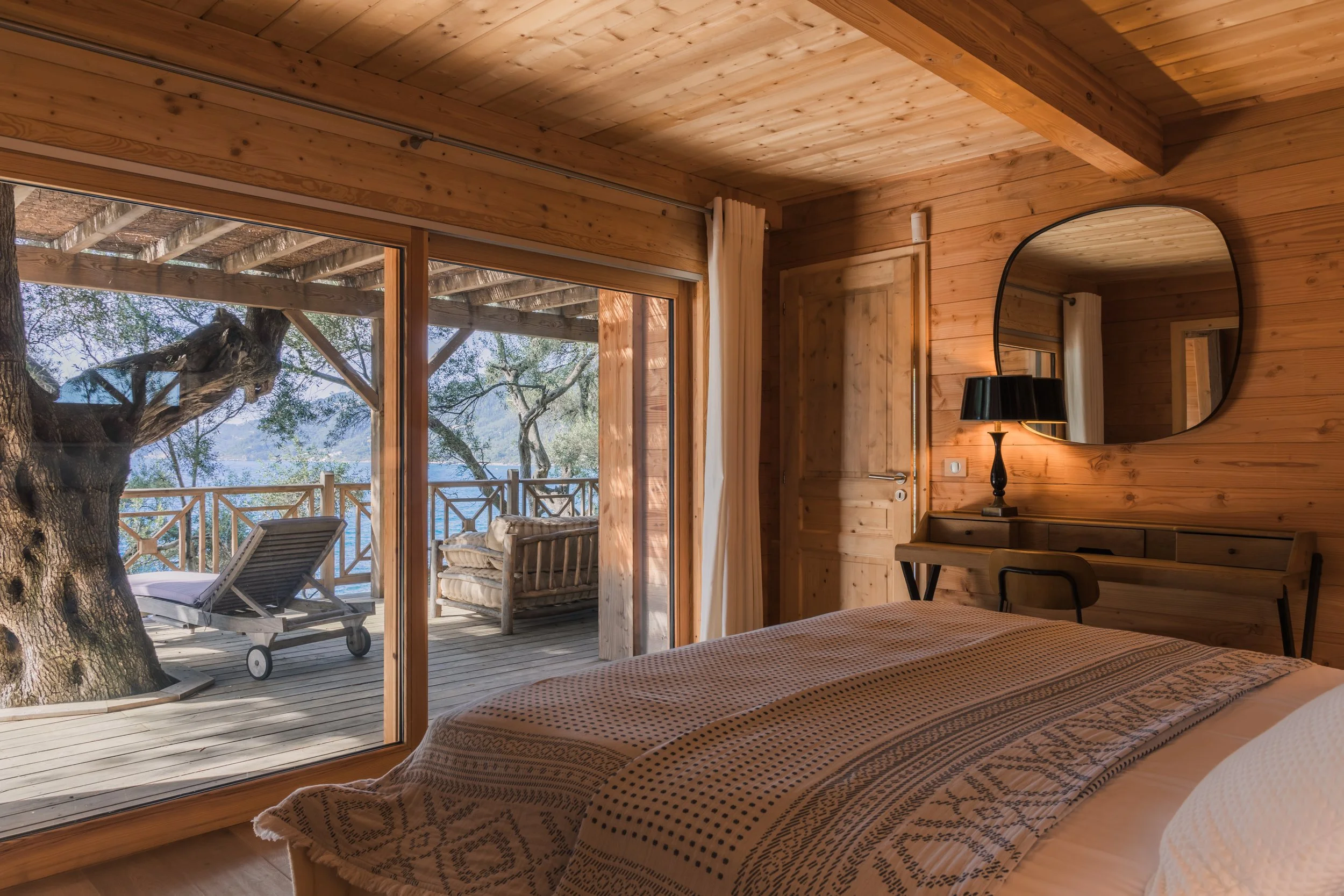 A cozy bedroom with wooden walls and ceiling, a bed with patterned blanket, a round mirror on the wall, a black lamp, and a desk with a chair. Outside, there is a wooden deck with outdoor furniture, a lounge chair, a sofa, surrounding trees, and a vi