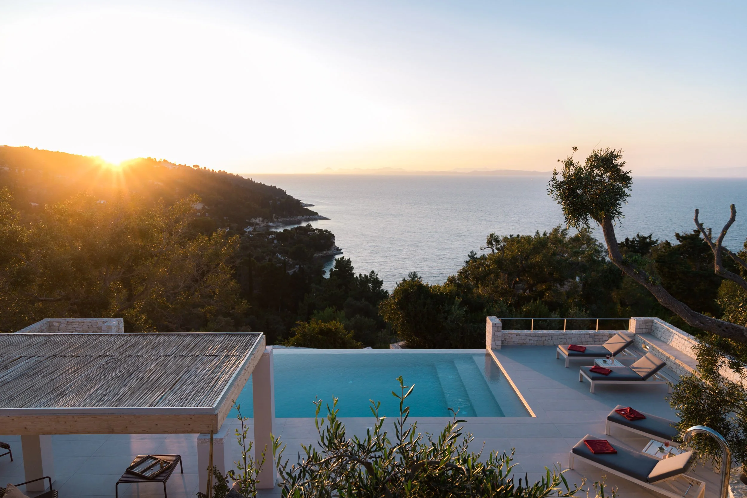 A scenic view of a sunset over the ocean, with trees and greenery in the foreground. In the lower part of the image, a swimming pool and four lounge chairs with red towels are visible on a terrace.