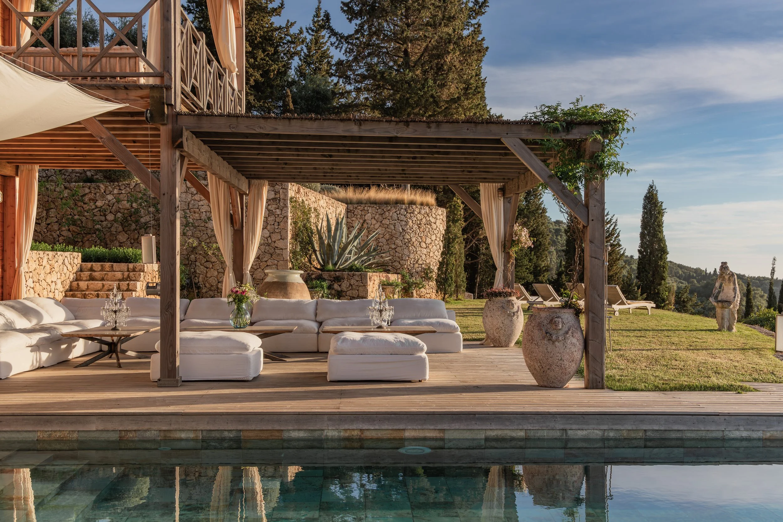 Outdoor patio with white sofa, glass tables, large vases, and surround landscaping including stone wall, agave plant, and trees, overlooking a landscape with hills and sky.
