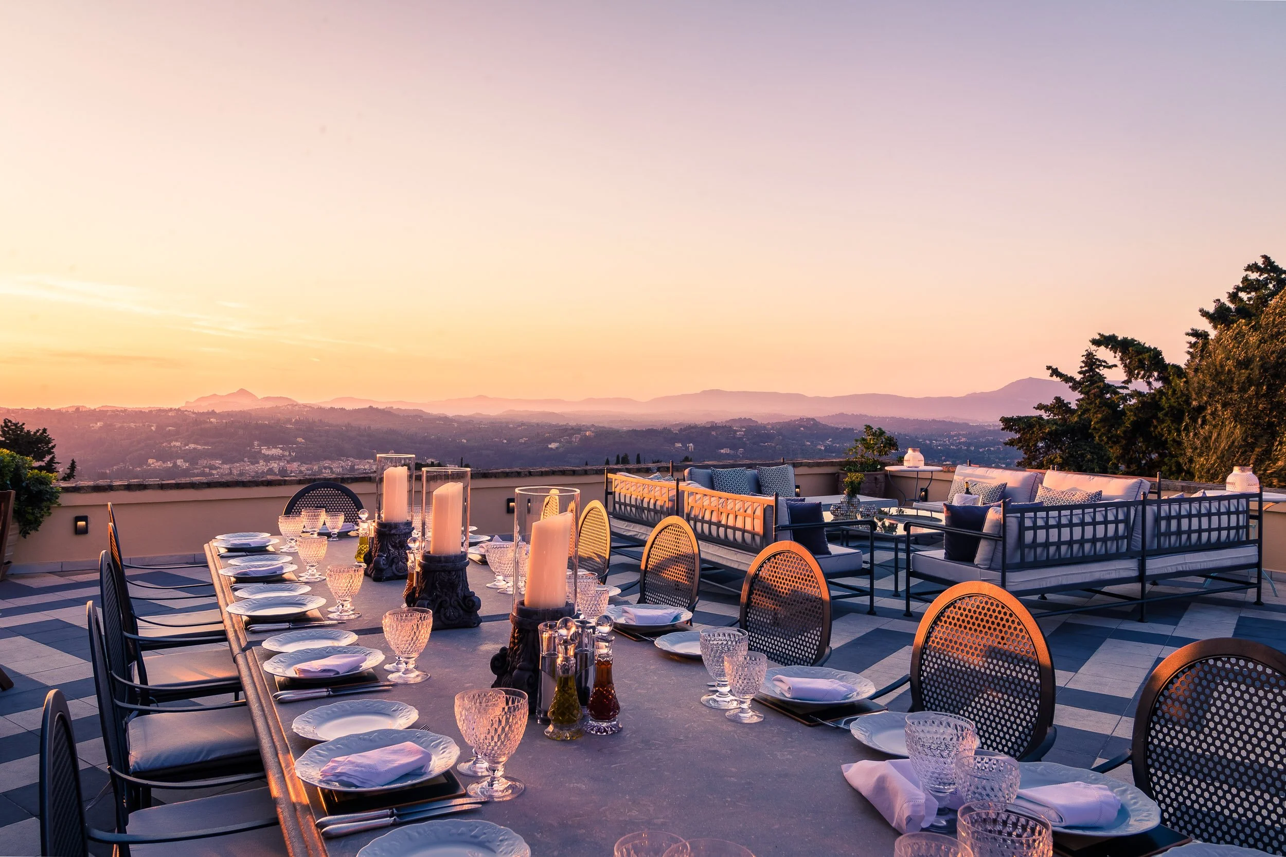 A rooftop dining area set for a meal with a long table, candles, and glassware, overlooking a cityscape at sunset with mountains in the distance.
