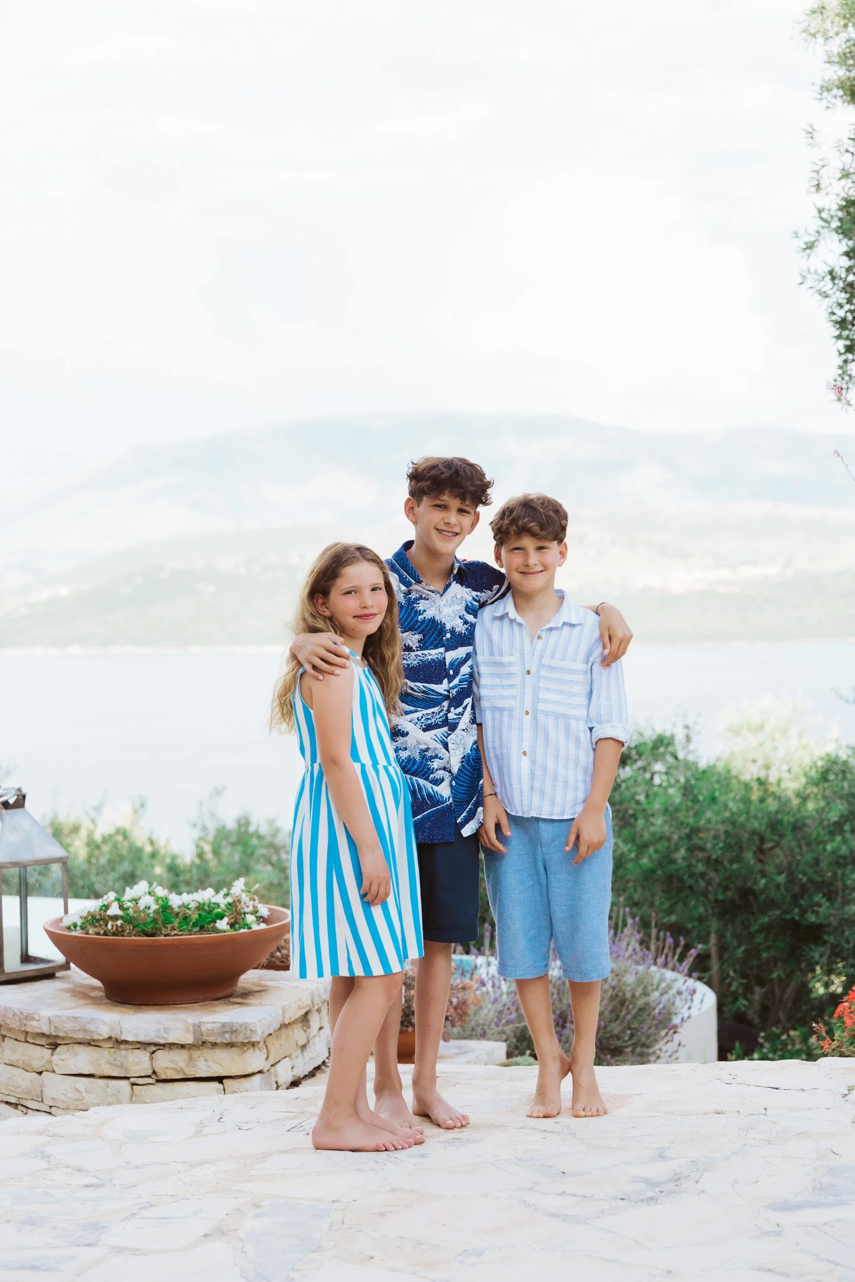 Three children standing outdoors near water, smiling, with a landscape of hills and trees in the background.