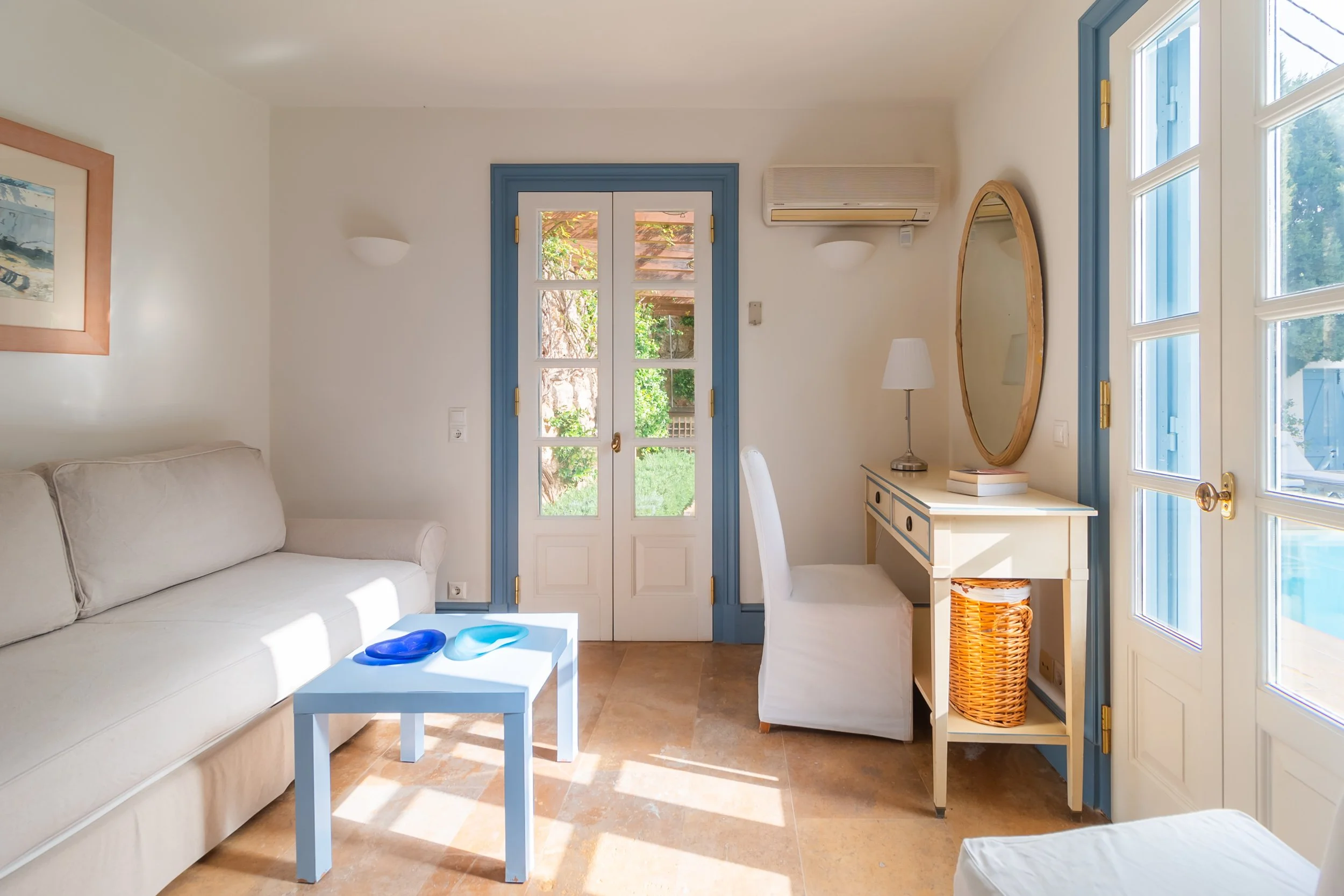 Bright living room with a beige sofa, a small blue coffee table with blue and turquoise plates, a white desk with a matching white chair, a round mirror, a table lamp, and a view of a sunny garden outside through blue and white French doors.