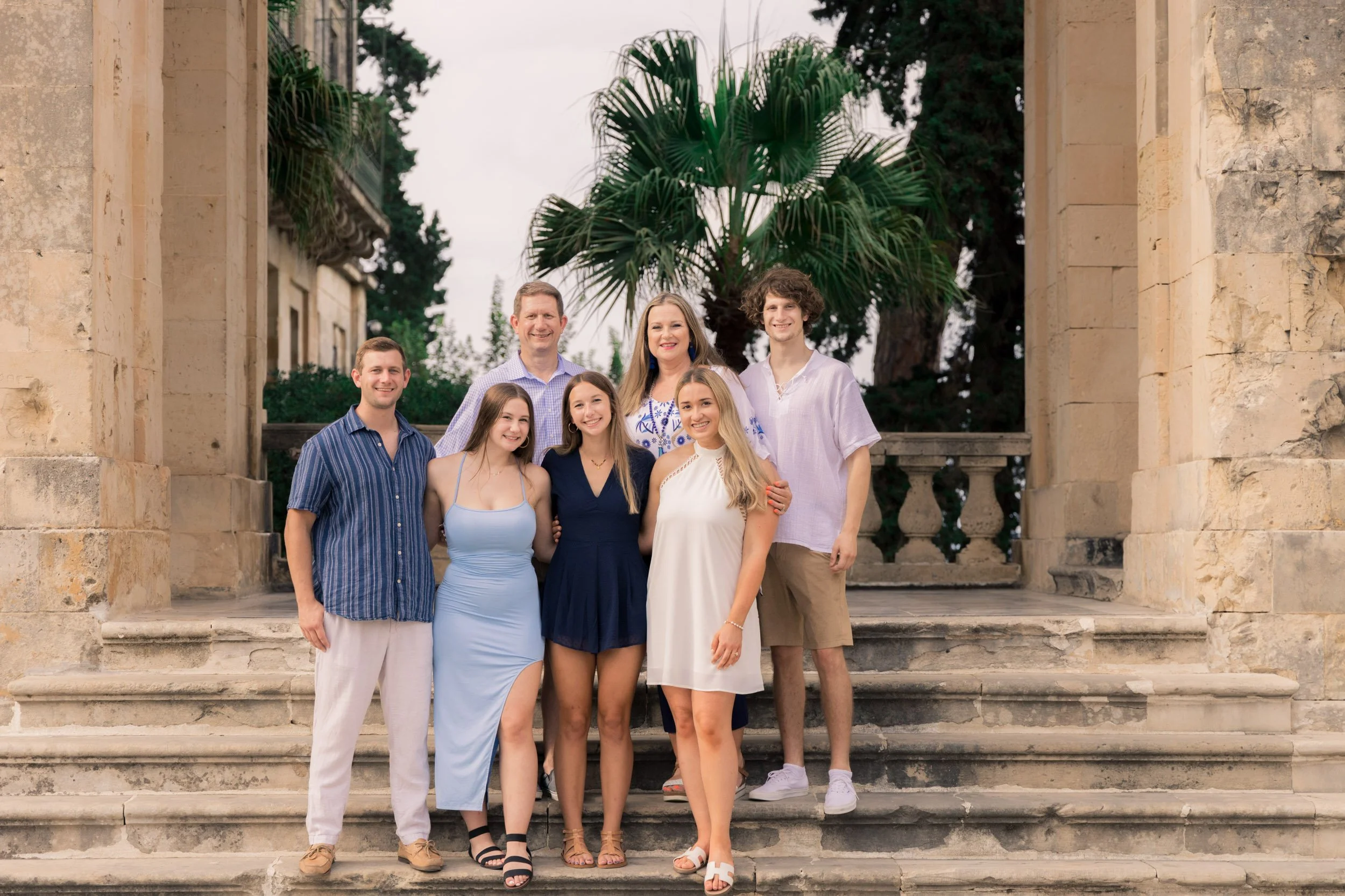 Group of eight people standing on stone steps in front of a palm tree and historic stone architecture