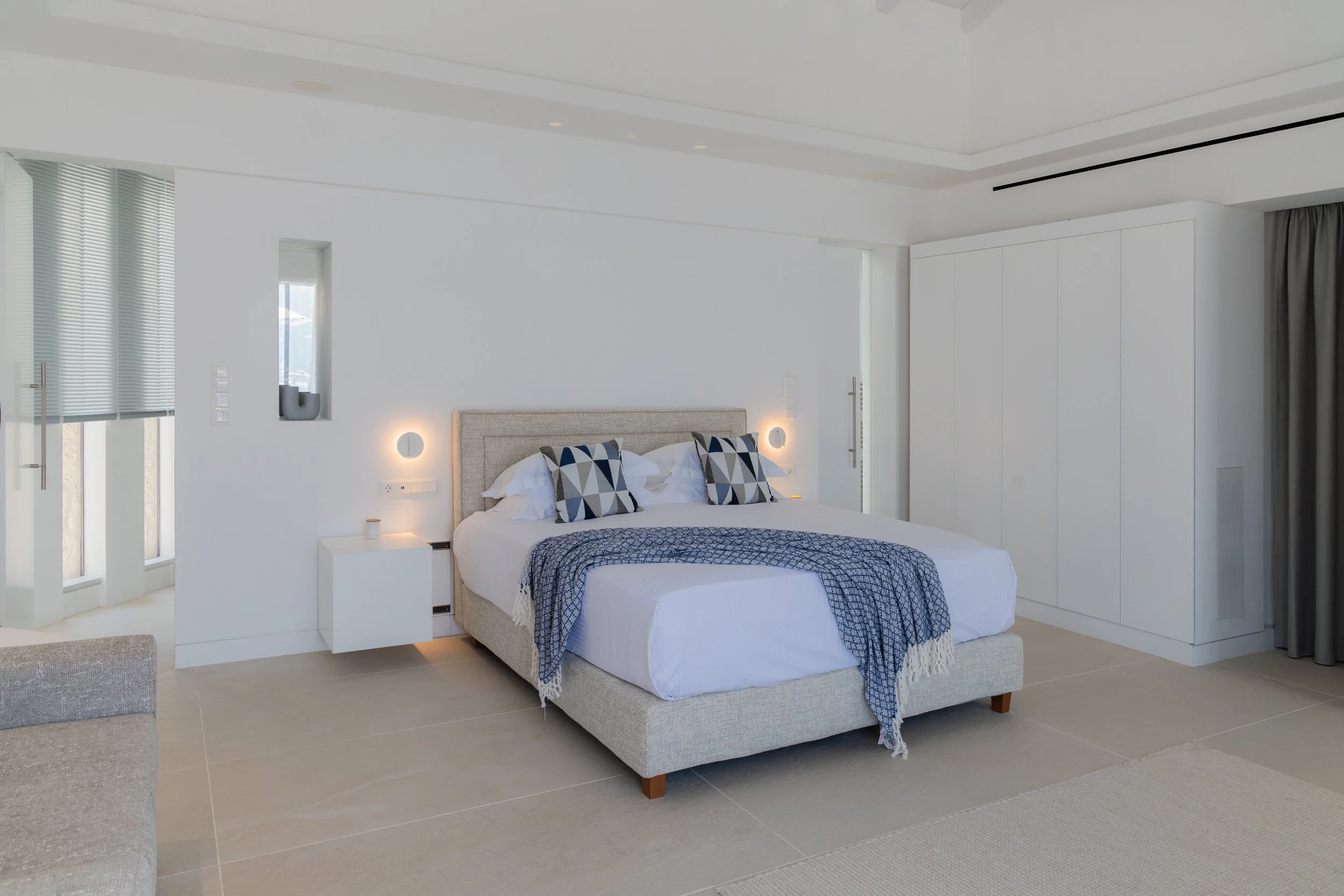 A neatly made bed with white bedding, blue patterned throw, and two geometric patterned pillows in a bright modern bedroom with beige tile floors, white walls, and large windows.