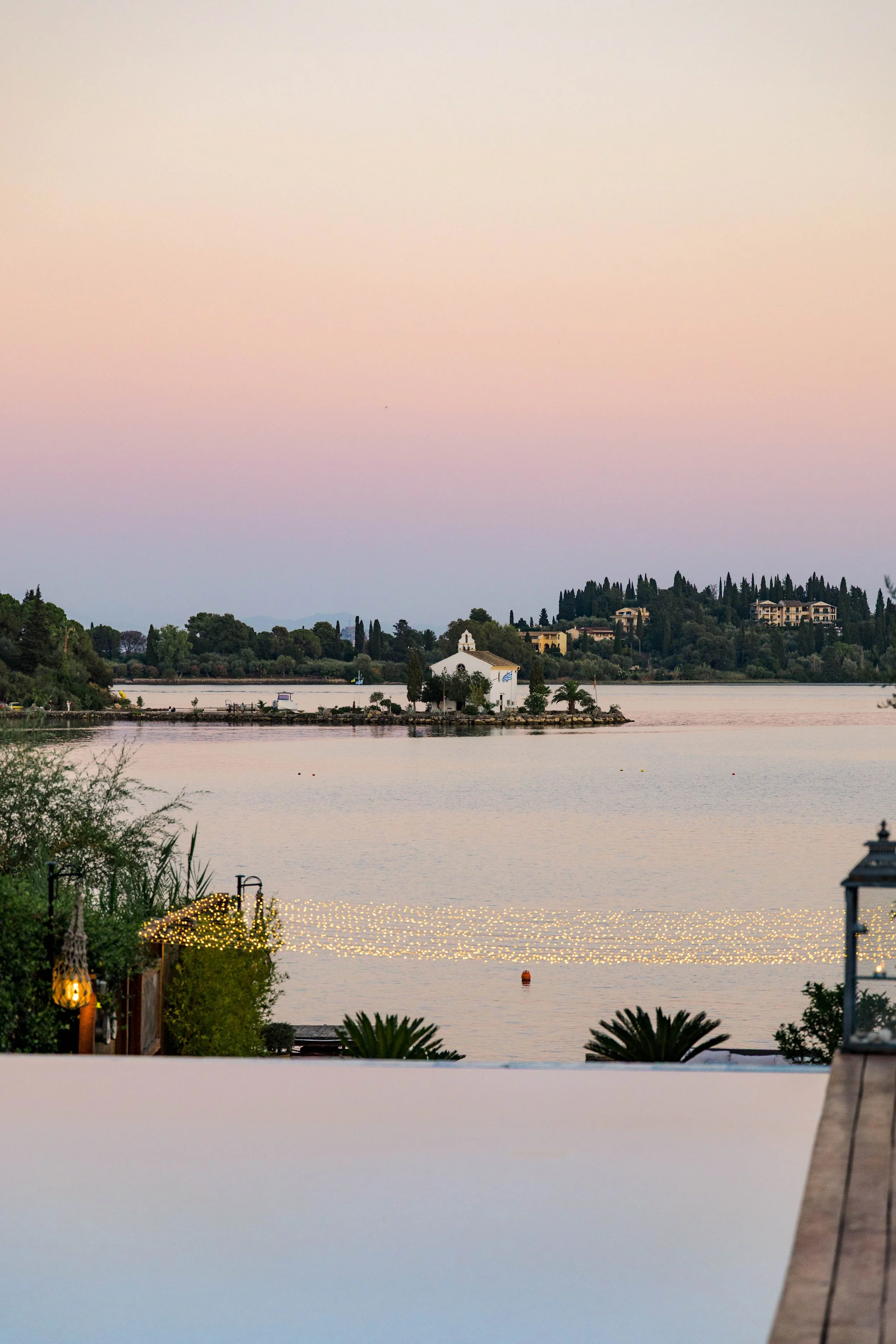 Scenic view of a lake at dusk, with a small white chapel on a peninsula in the middle, trees on the shore, and buildings in the background. Decorative lights are strung along the shoreline.