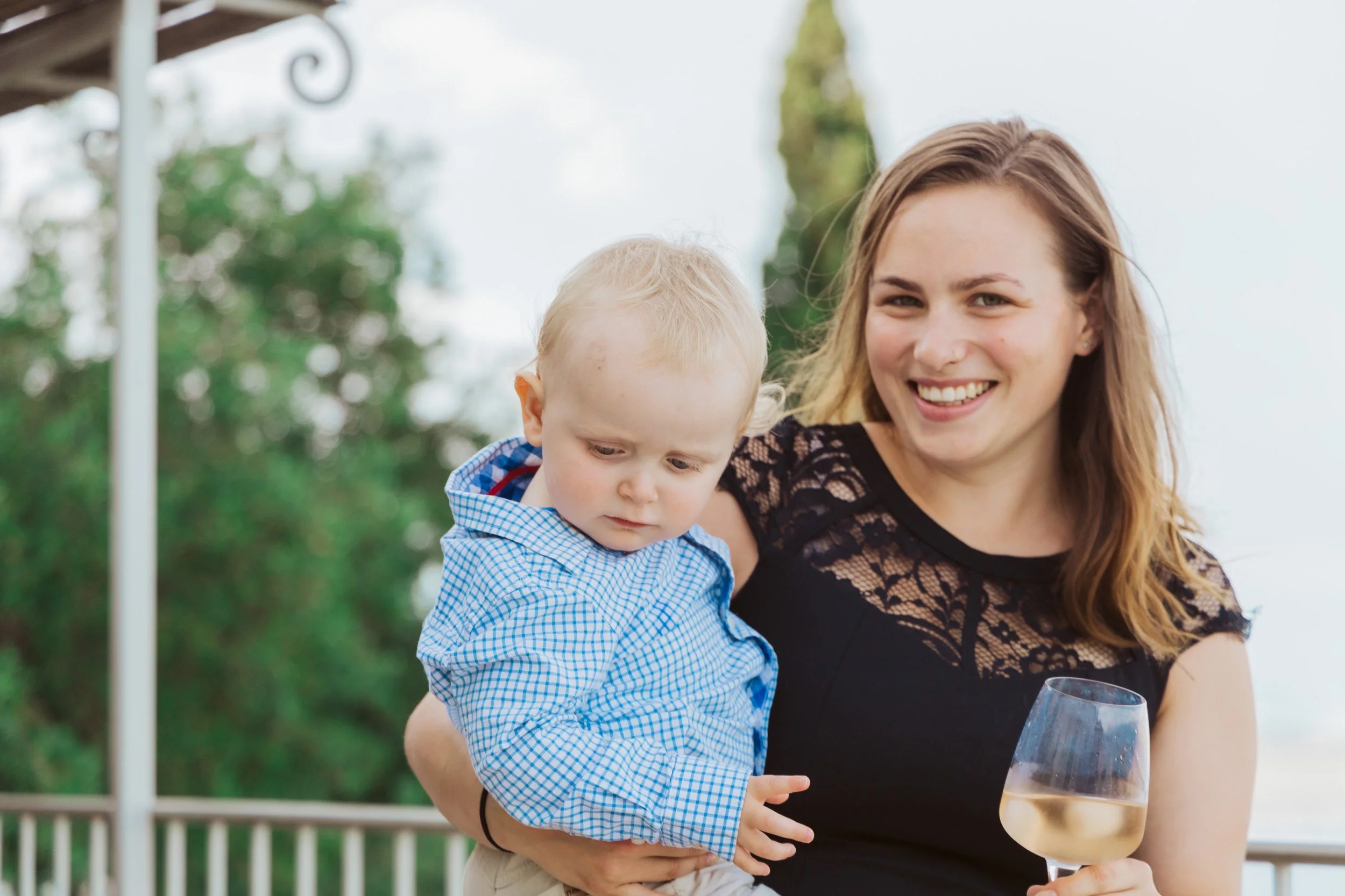 A woman holding a young child outdoors on a balcony, smiling while holding a glass of white wine, with trees and a cloudy sky in the background.