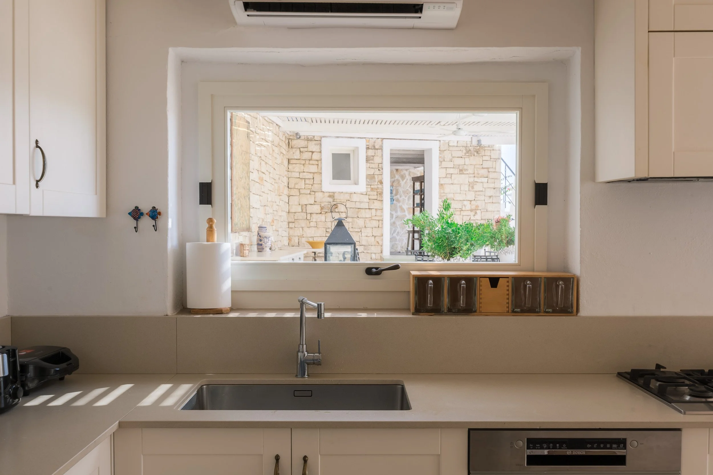 View from a kitchen sink looking out a window to a patio with stone wall, potted plants, and outdoor furniture.