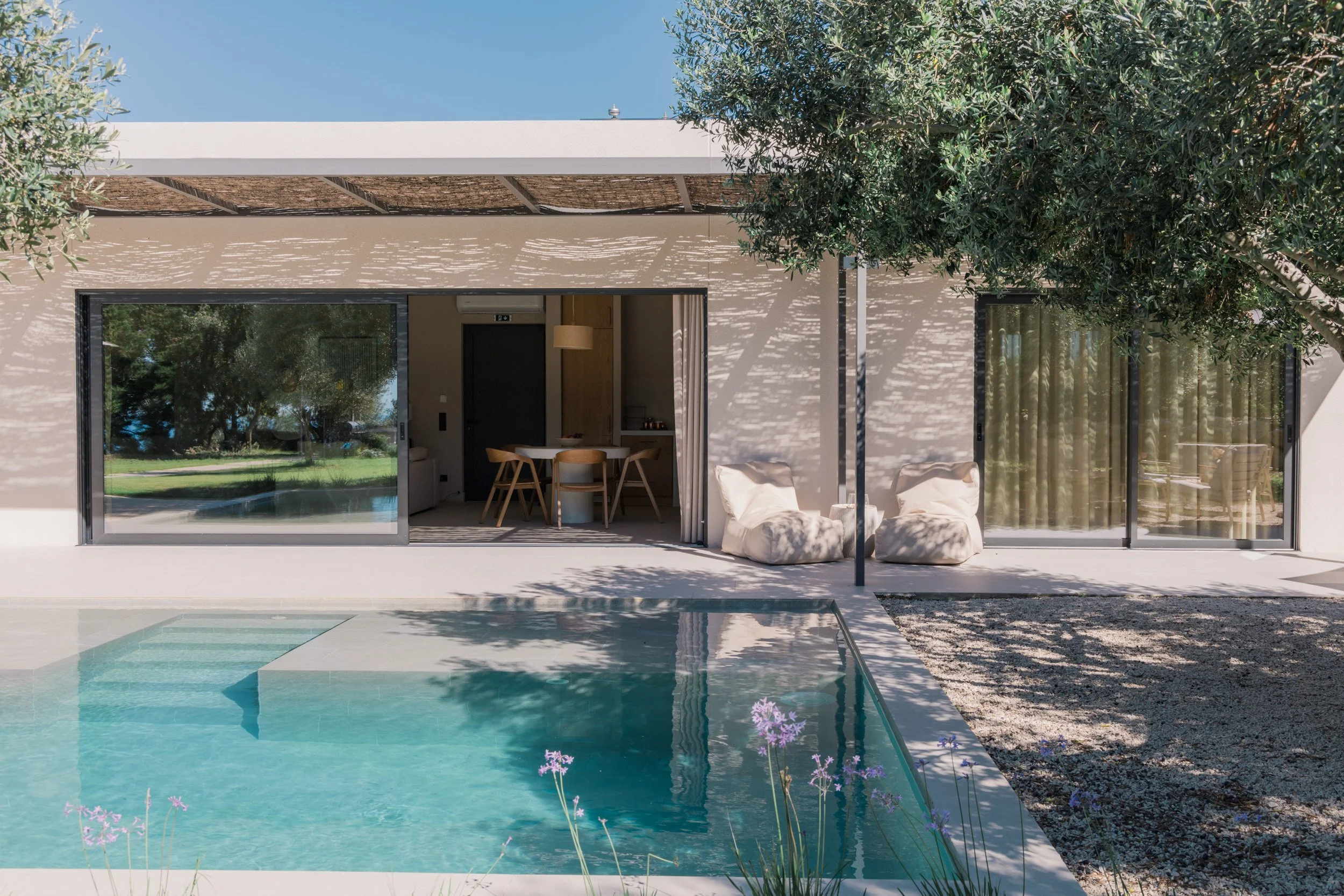 Modern house with large sliding glass doors, outdoor sitting area with bean bag chairs, a swimming pool, and a backyard with trees and grass.