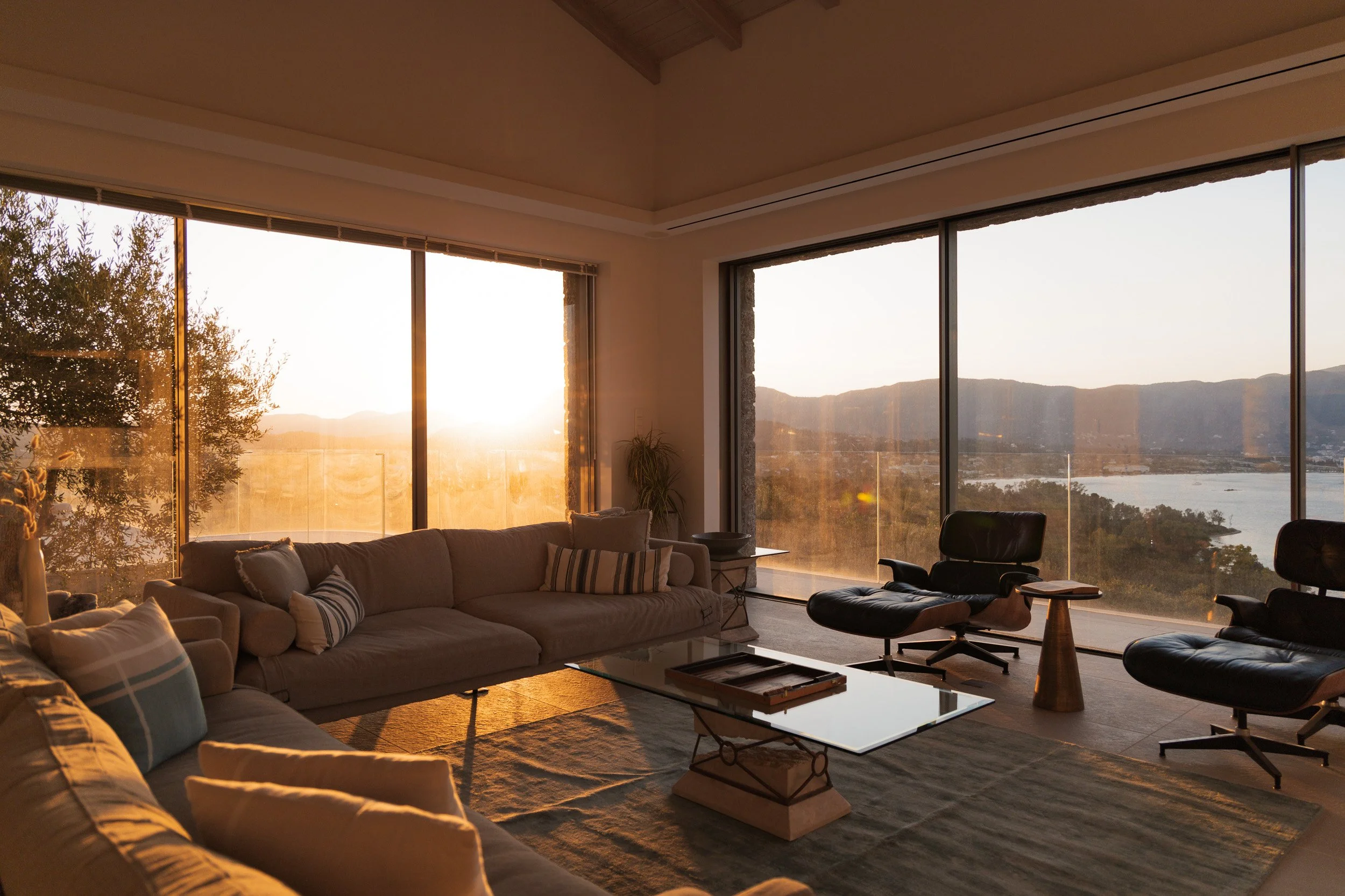 Living room with large floor-to-ceiling windows showing a sunset over a lake and mountains, with sofas, a glass-top coffee table, and lounge chairs.