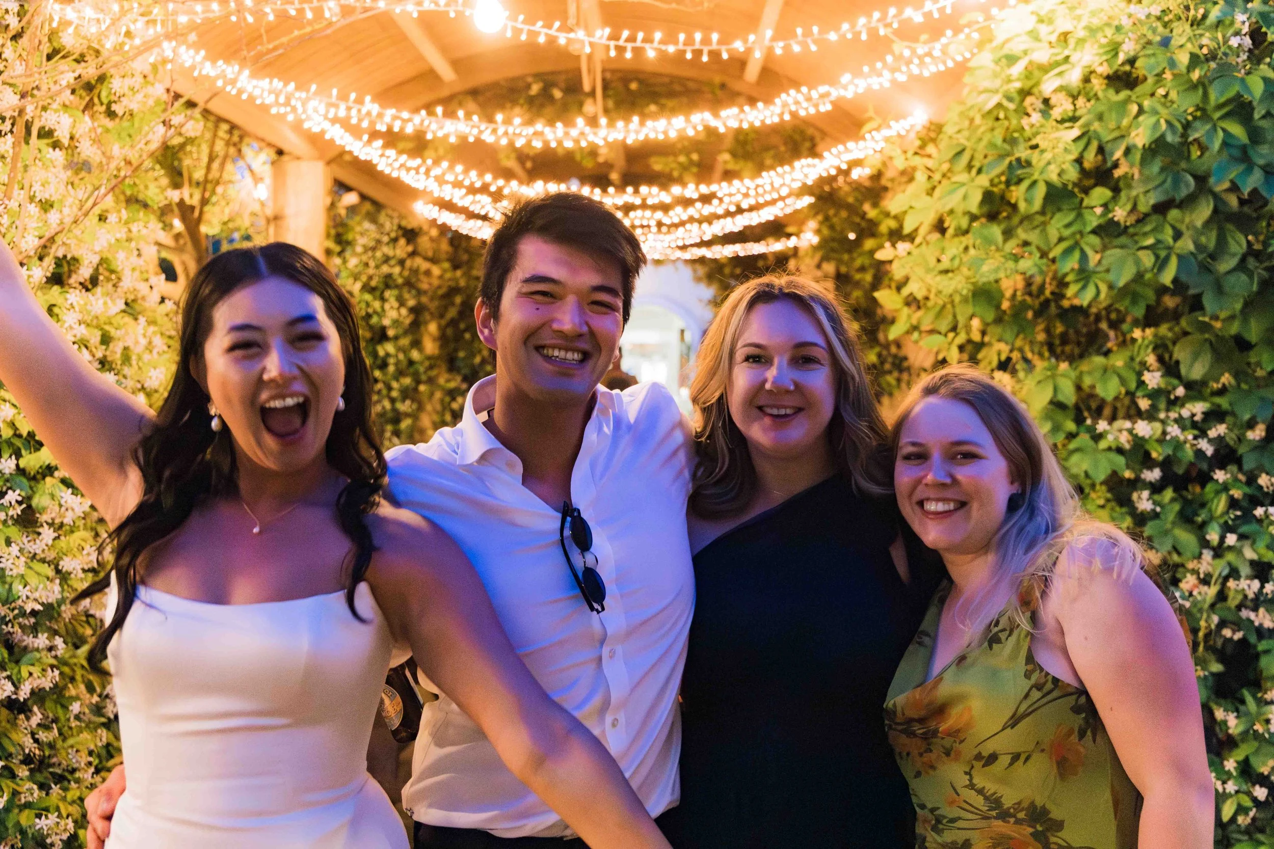 Four young adults smiling and celebrating under string lights adorned with greenery and white flowers.