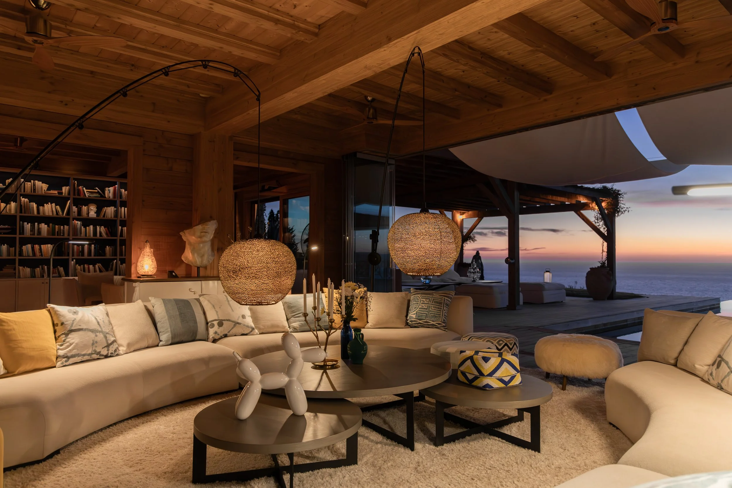 Living room with a large white sectional sofa, abstract throw pillows, a round coffee table with vases and decorative items, textured pendant lights, a bookcase in the background, and an outdoor view of a sunset over the ocean.