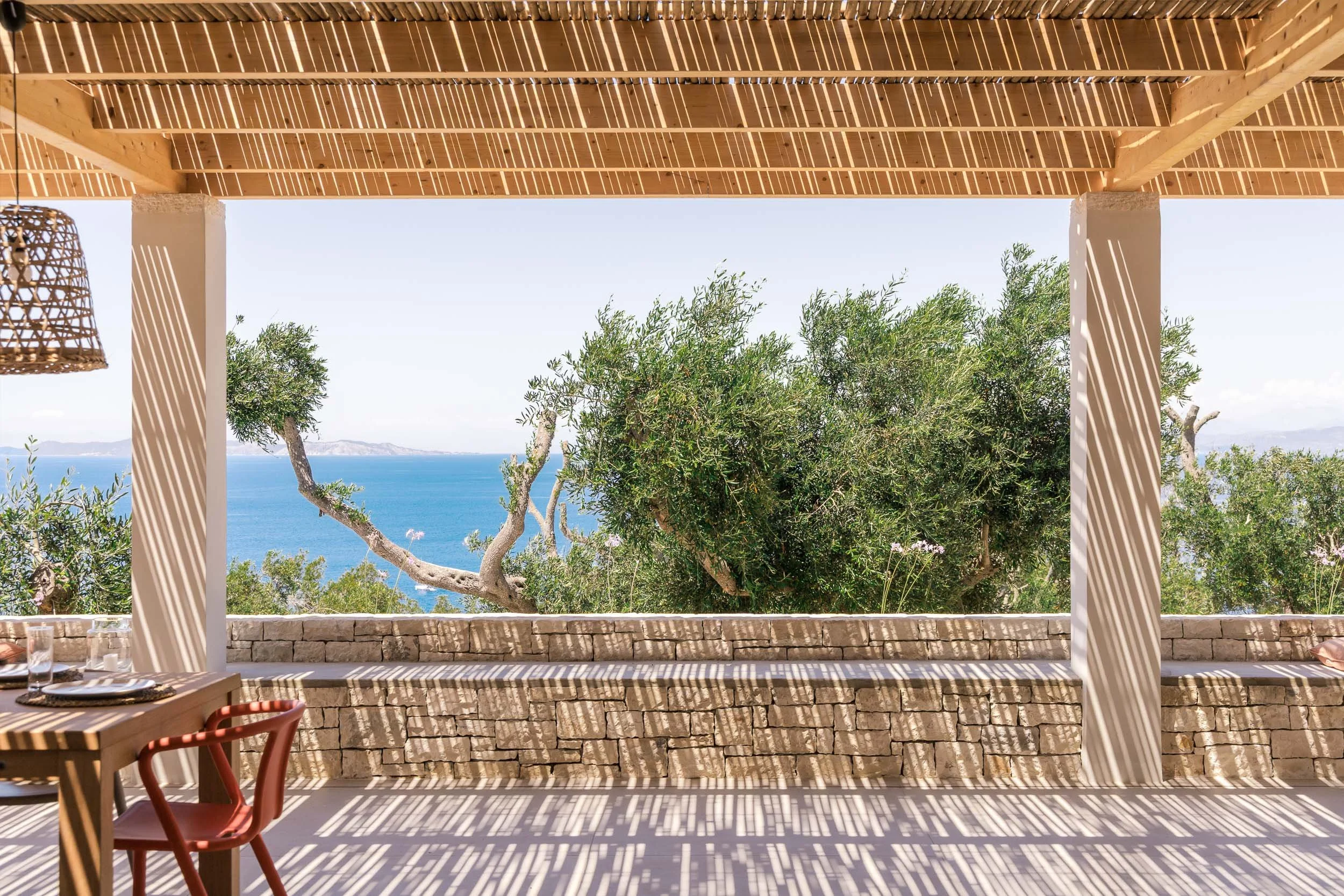 View from a covered patio with a stone wall, overlooking trees and the ocean in the distance on a sunny day.