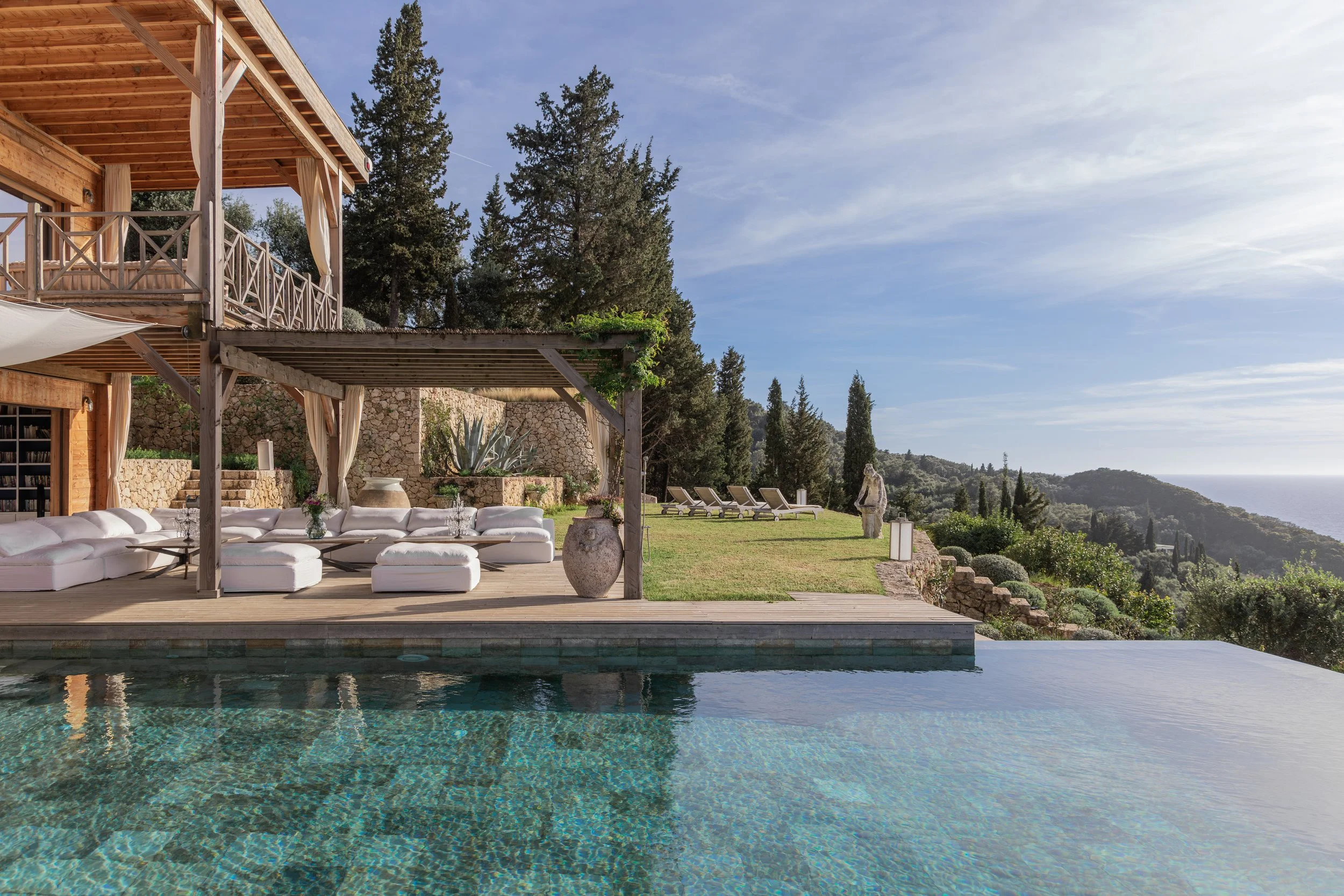 Luxury outdoor setting with a swimming pool, white lounge furniture under a wooden deck, garden chairs on the grass, surrounded by trees and a scenic view of hills and the ocean in the distance.