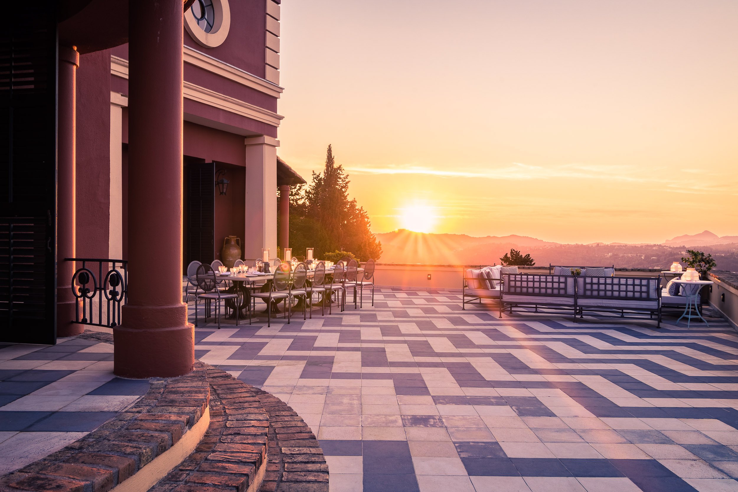 A spacious outdoor terrace with a checkered tile floor, decorated with tables and chairs, overlooking a sunrise or sunset over distant hills.