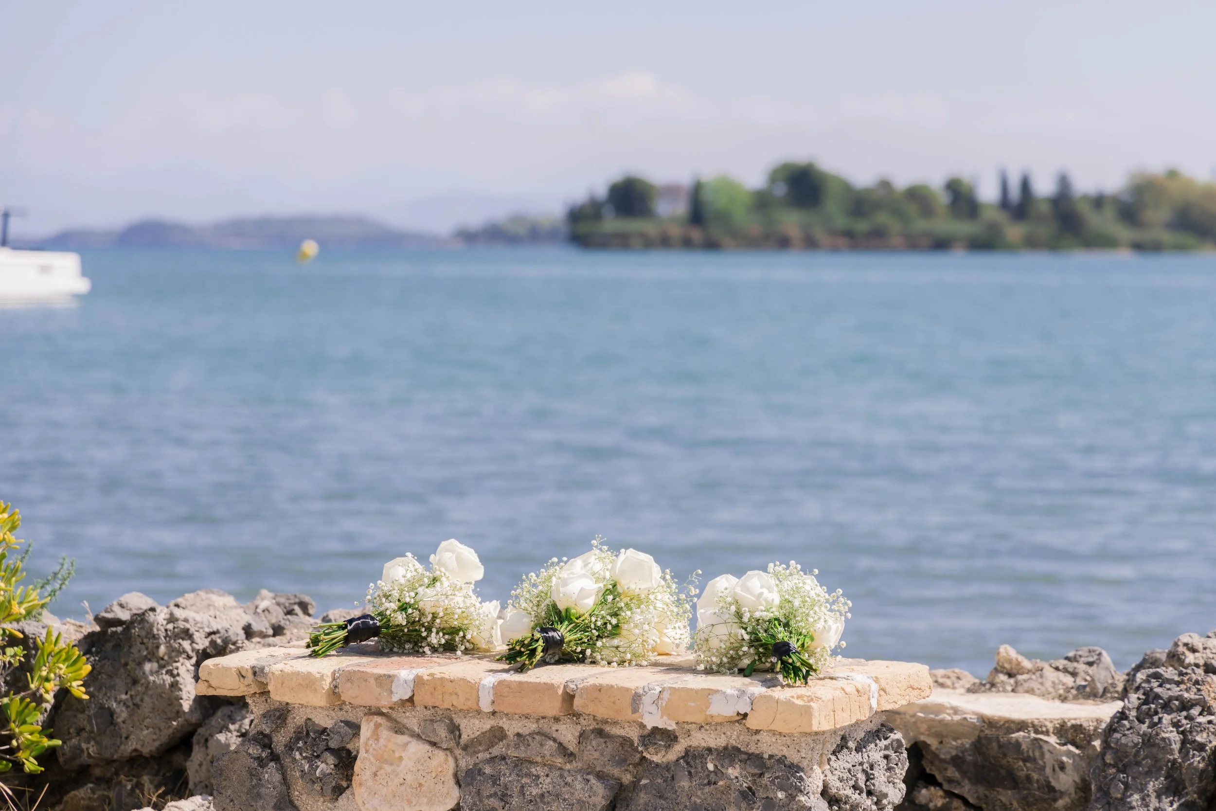 Three small floral bouquets with white roses and baby's breath arranged on a stone ledge by the water, with an island and a boat in the background under a partly cloudy sky.