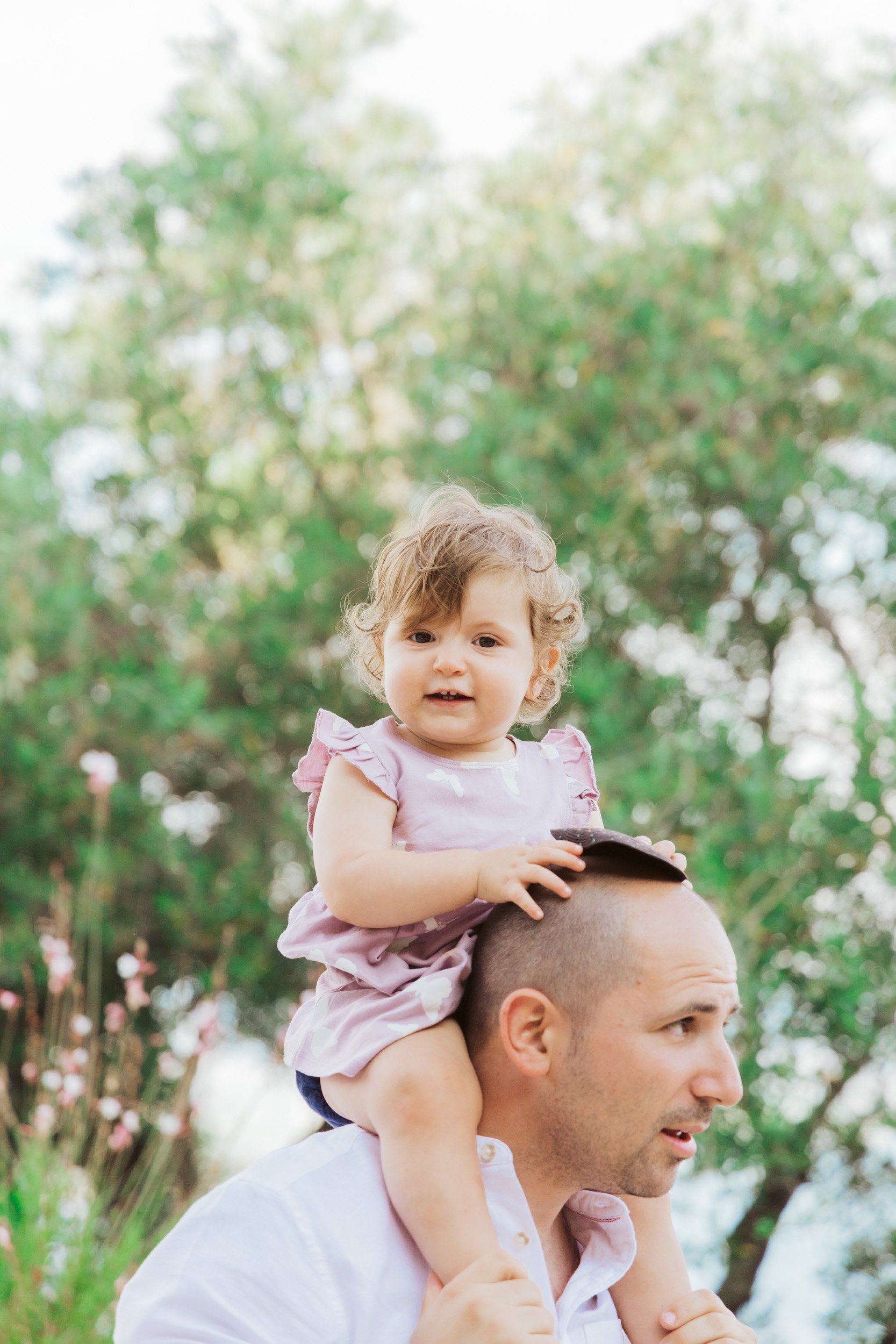 A young girl with curly hair sitting on a man's shoulders outdoors in front of green trees.