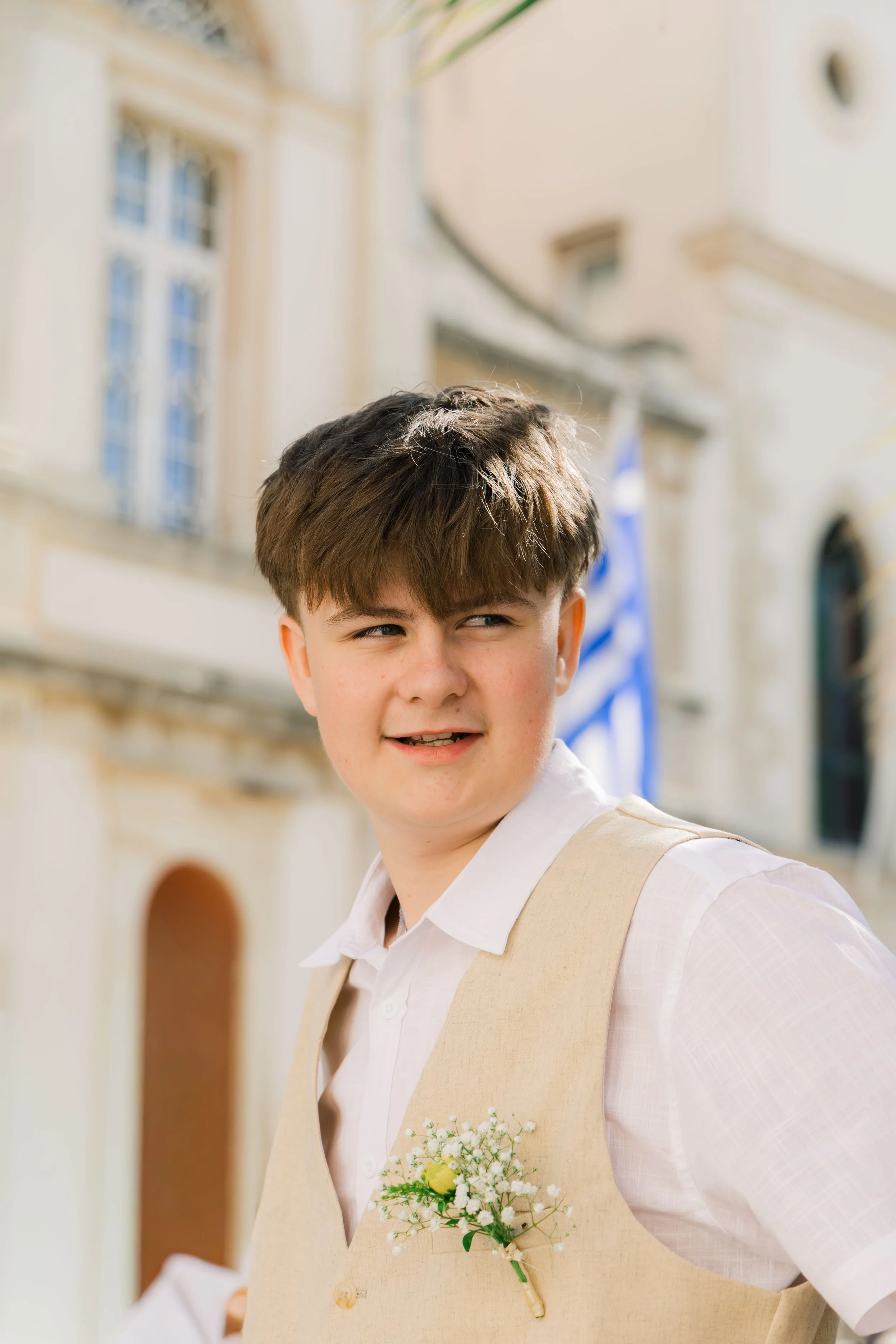 Young boy outdoors in formal attire with a beige vest and white shirt, wearing a small boutonniere, standing near a building with Greek flag in background.
