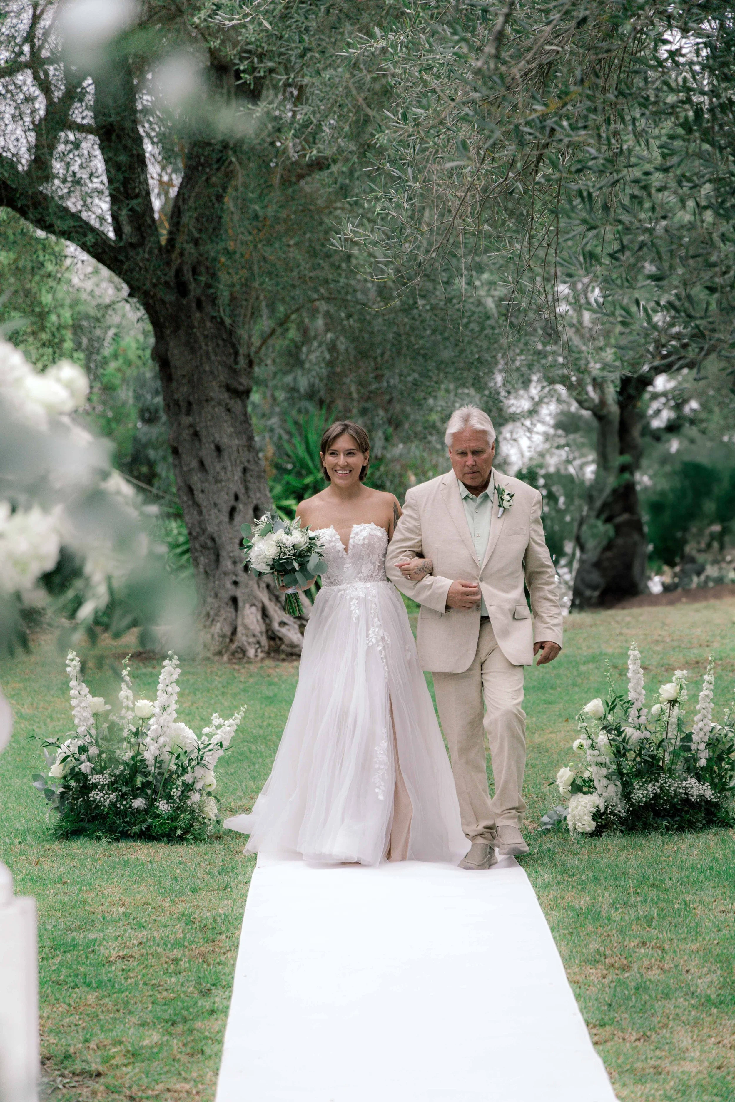 A bride holds a bouquet of white and green flowers, walking arm-in-arm with an older man in a beige suit during a wedding ceremony outdoors under trees, on a white aisle runner decorated with floral arrangements.