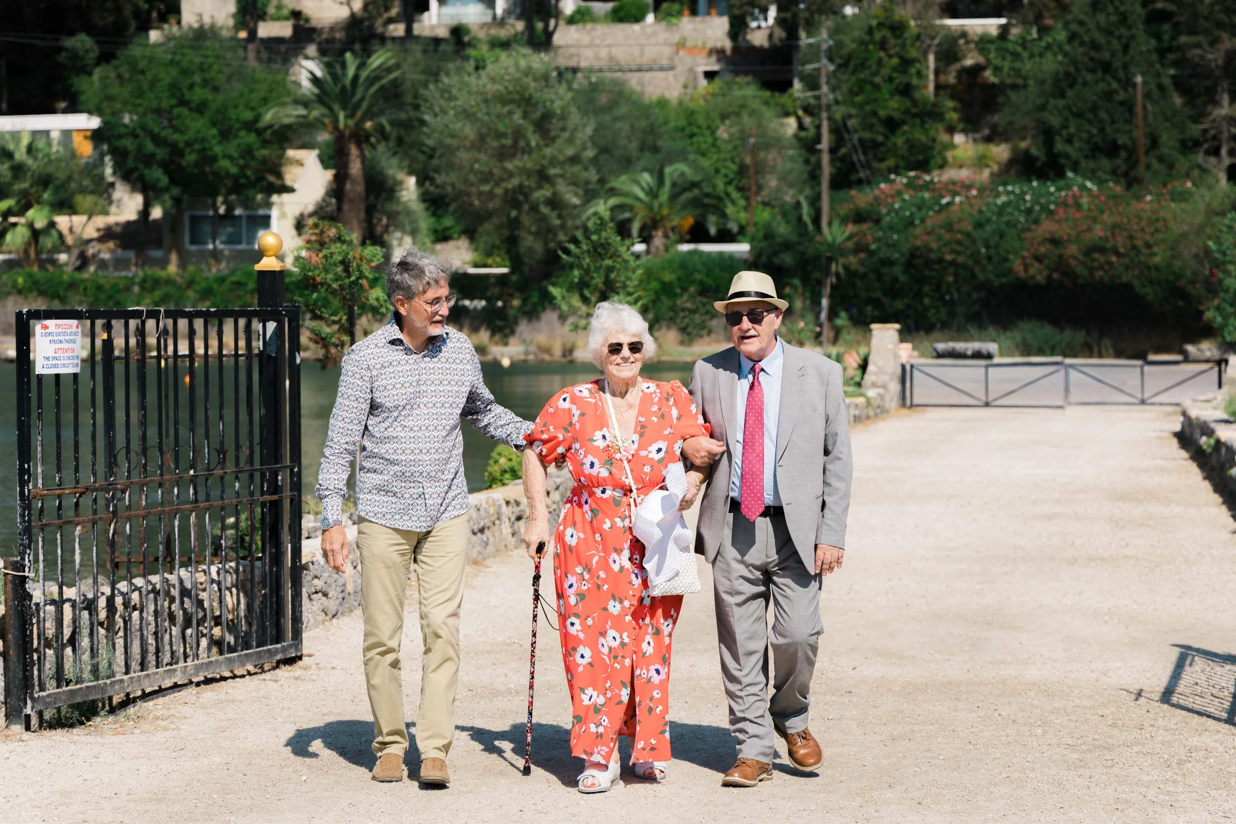 Three elderly people walking arm in arm outdoors on a sunny day, near a body of water with green trees and a stone wall in the background. One woman is wearing a vibrant orange floral dress, and two men are dressed in casual and semi-formal attire.