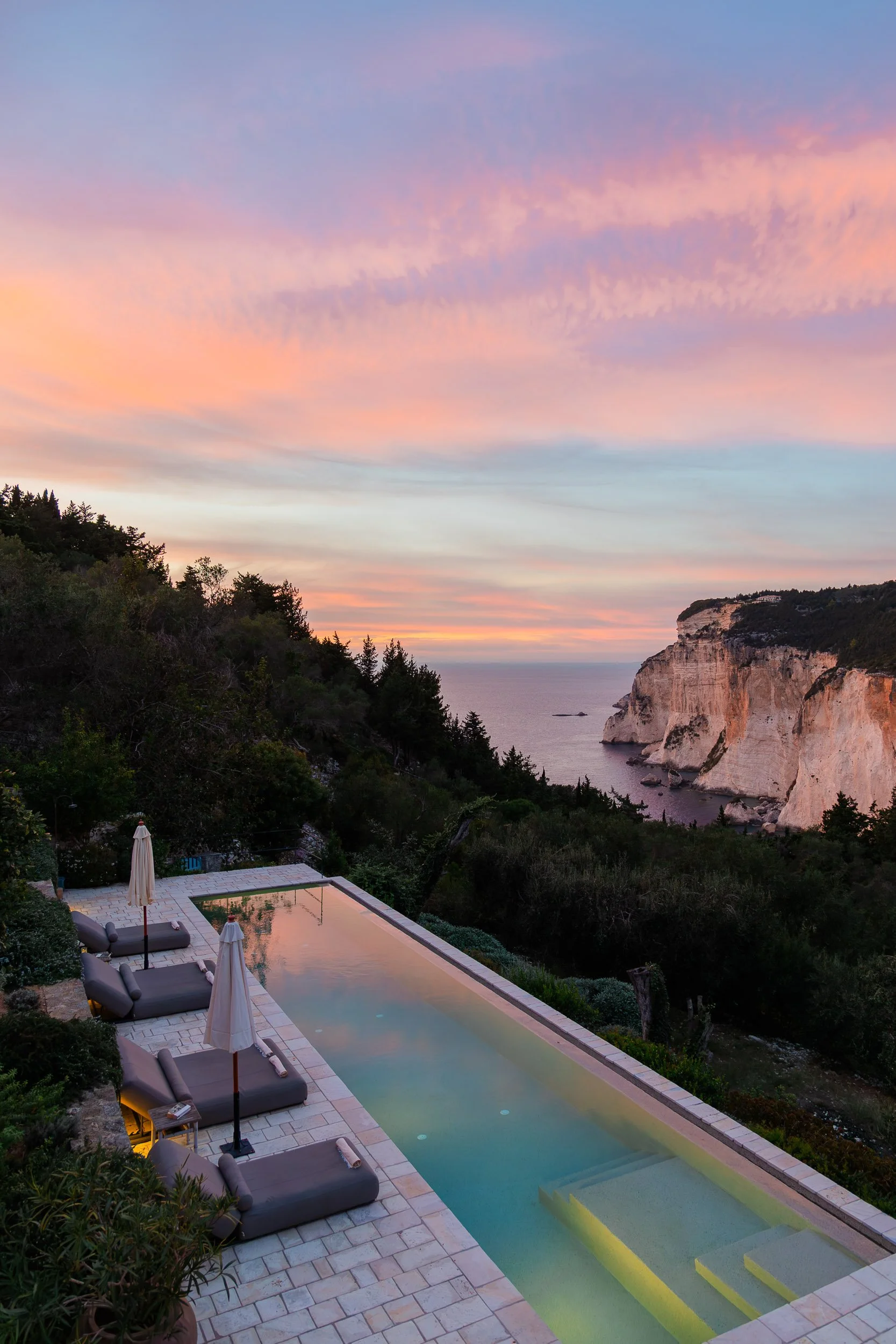 An infinity pool overlooking a coastal cliff at sunset with pastel-colored sky and calm ocean water.