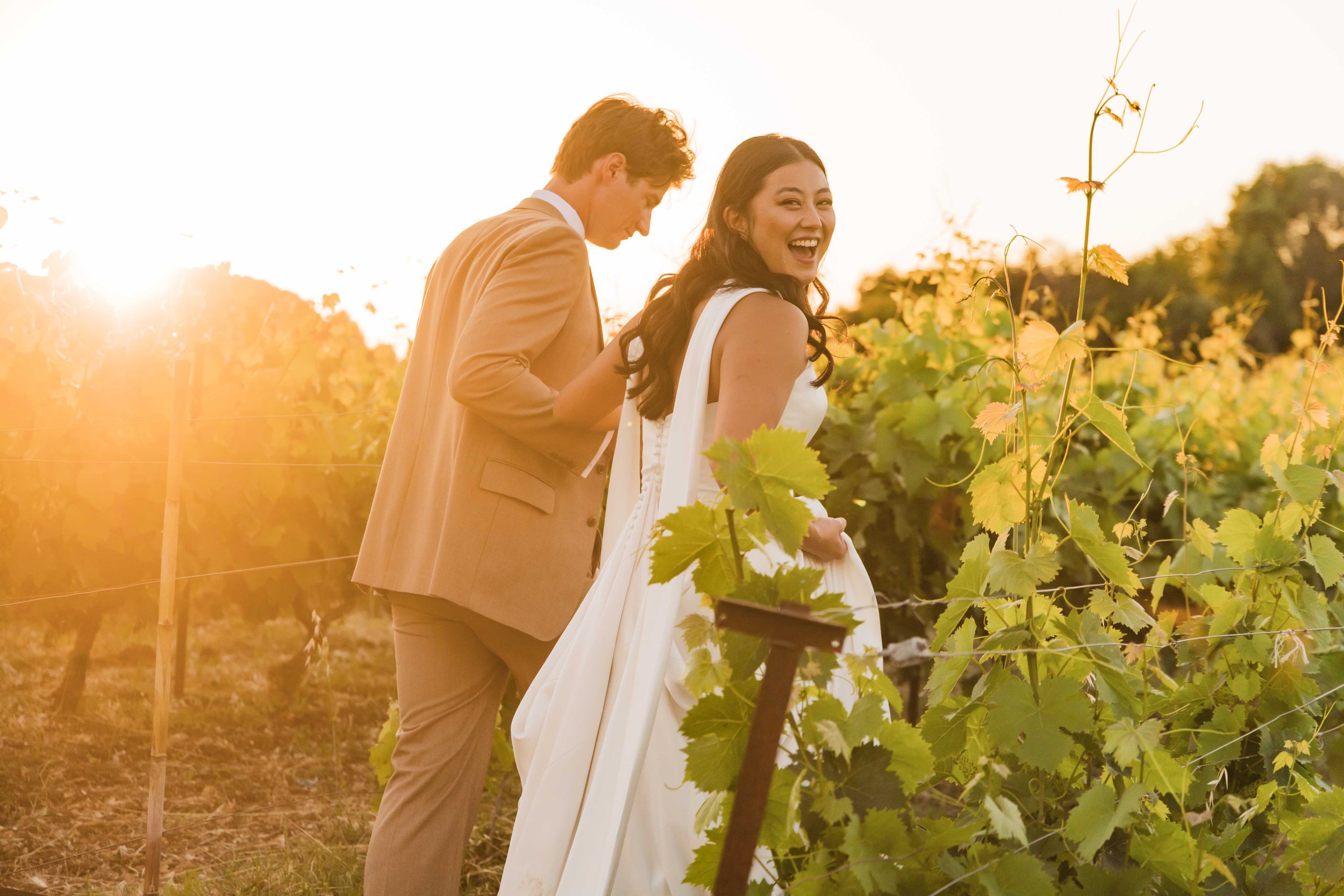 A happy couple in wedding attire standing in a vineyard at sunset.