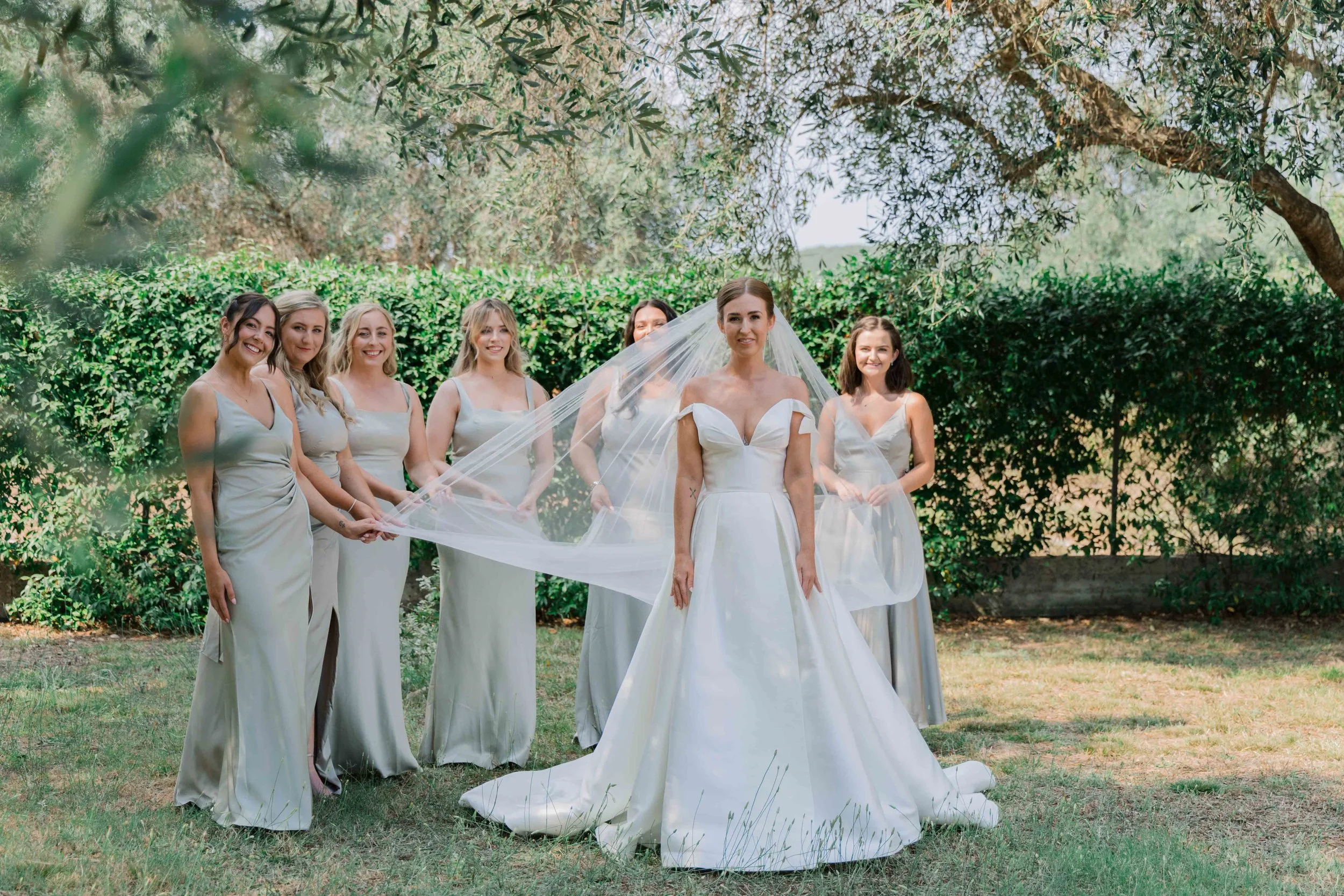A bride in a white wedding gown and veil standing outdoors with six bridesmaids in matching silver dresses.