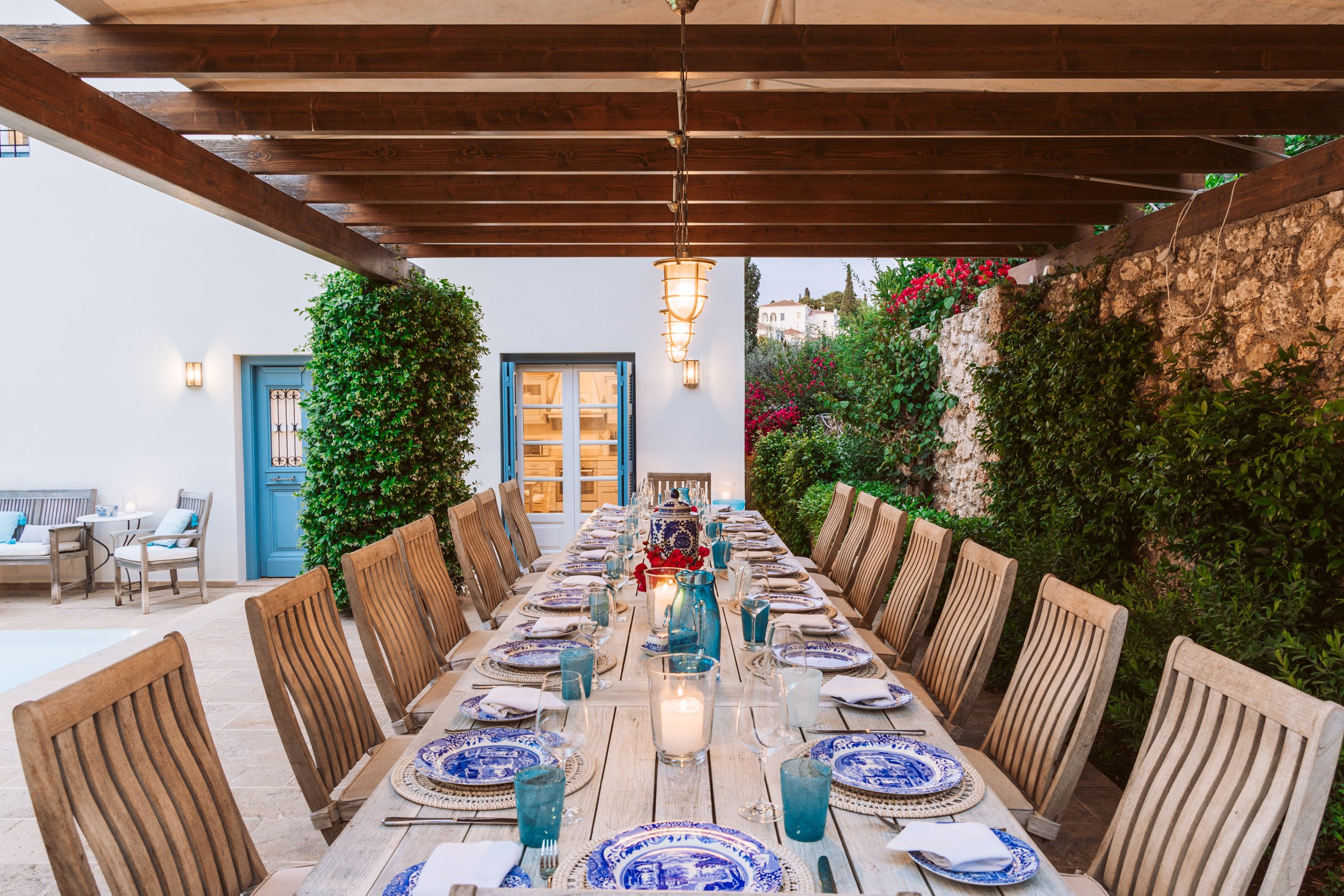 Outdoor dining area with a long wooden table set with blue and white plates, glasses, silverware, and candles, beneath a wooden pergola, with chairs around the table, greenery along the wall, and a white building with blue door and shutters in the ba