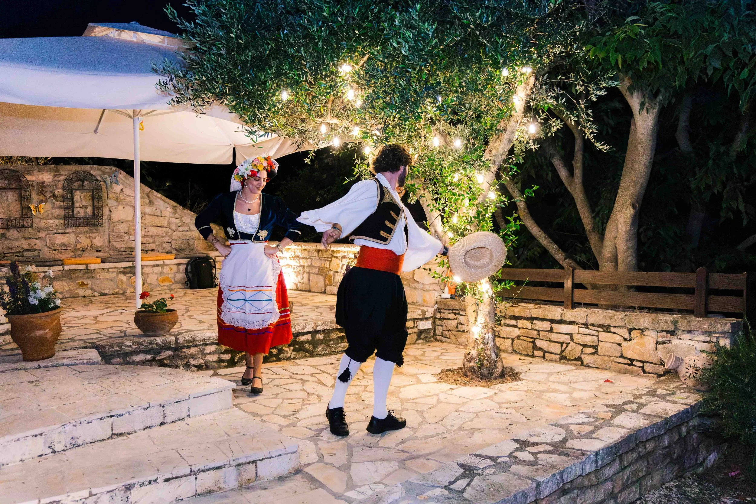 Two people dressed in traditional folk costumes performing a dance outdoors at night, illuminated by string lights, with trees and plants around and a stone patio surface.