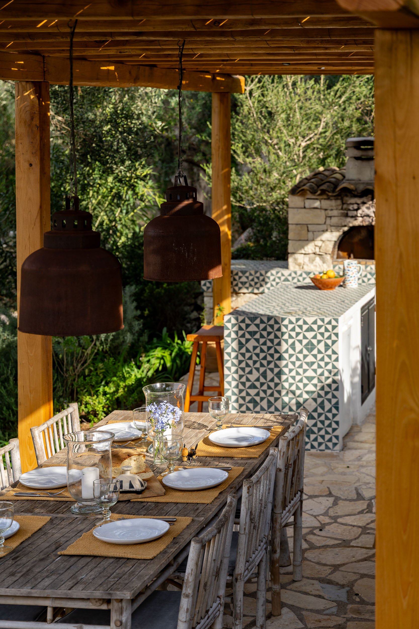Outdoor dining area with a rustic wooden table set with plates, glasses, and a small floral centerpiece, under hanging black lamps, with a patterned outdoor kitchen and greenery in the background.