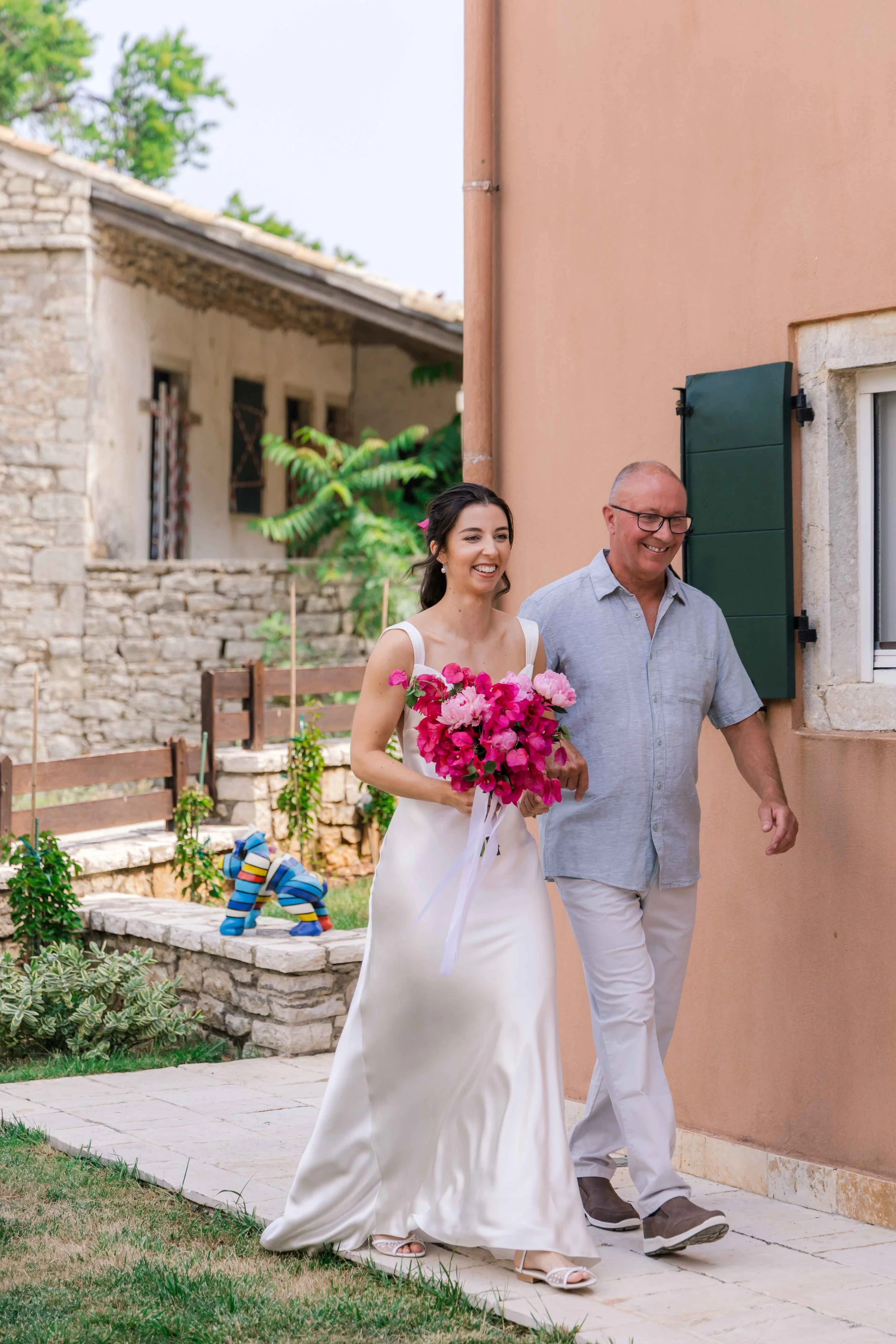 A woman in a white wedding dress holding a pink bouquet walks with an older man in a gray shirt and white pants outside a peach-colored house with green shutters, smiling.