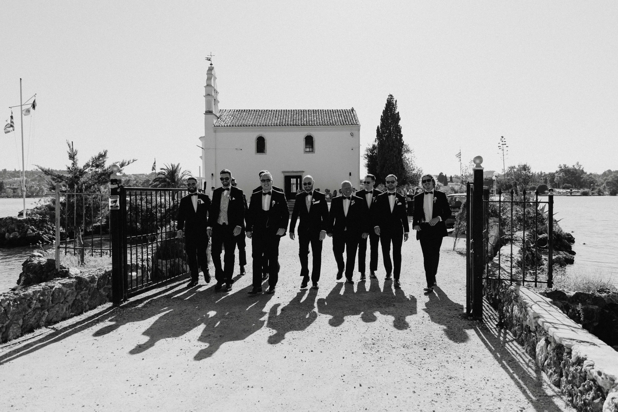 A group of men in tuxedos walking through an open gate, casting long shadows, with a small white chapel and trees in the background, near a body of water.