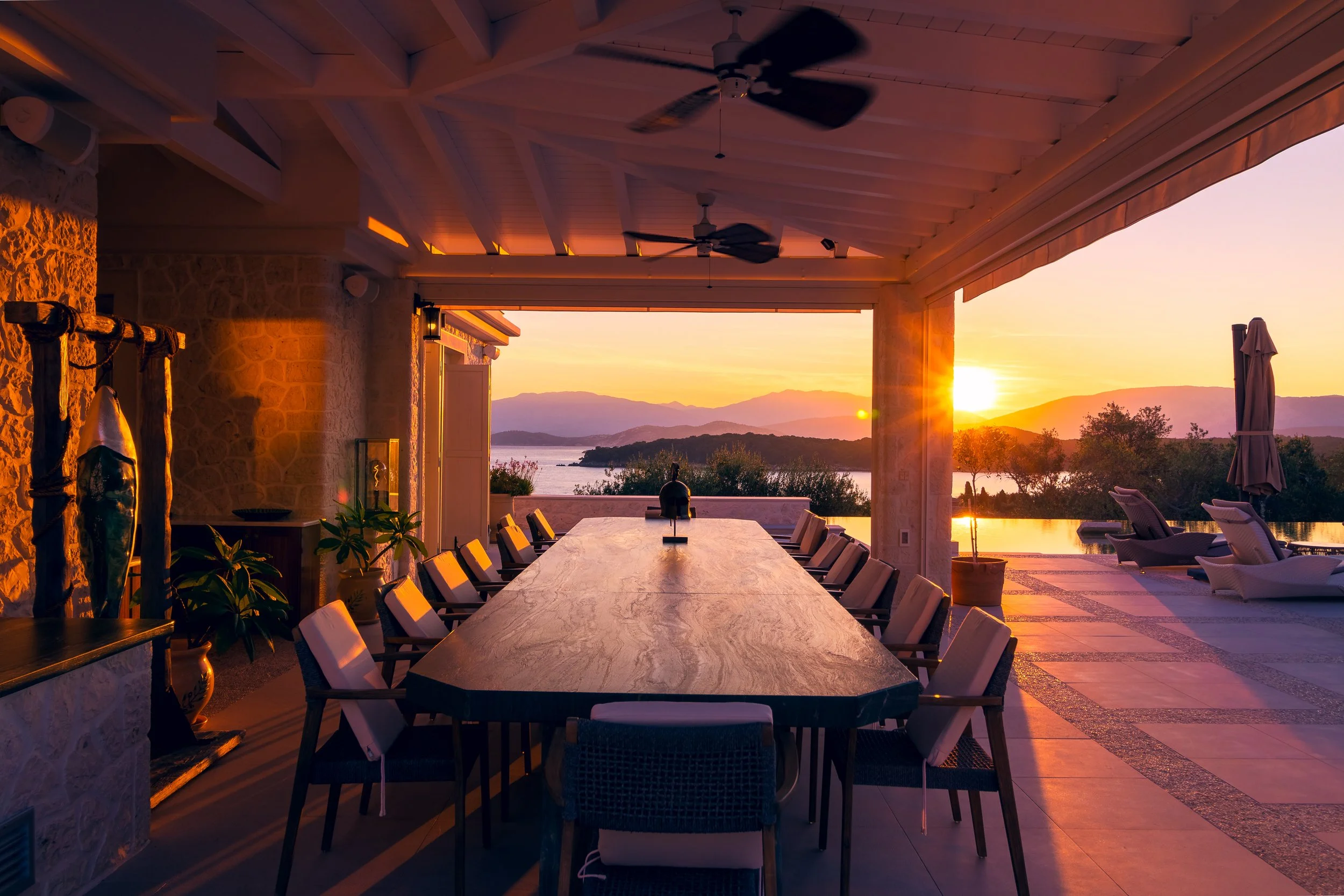 A covered outdoor dining area with a large table and chairs, overlooking a lake and mountains at sunset.