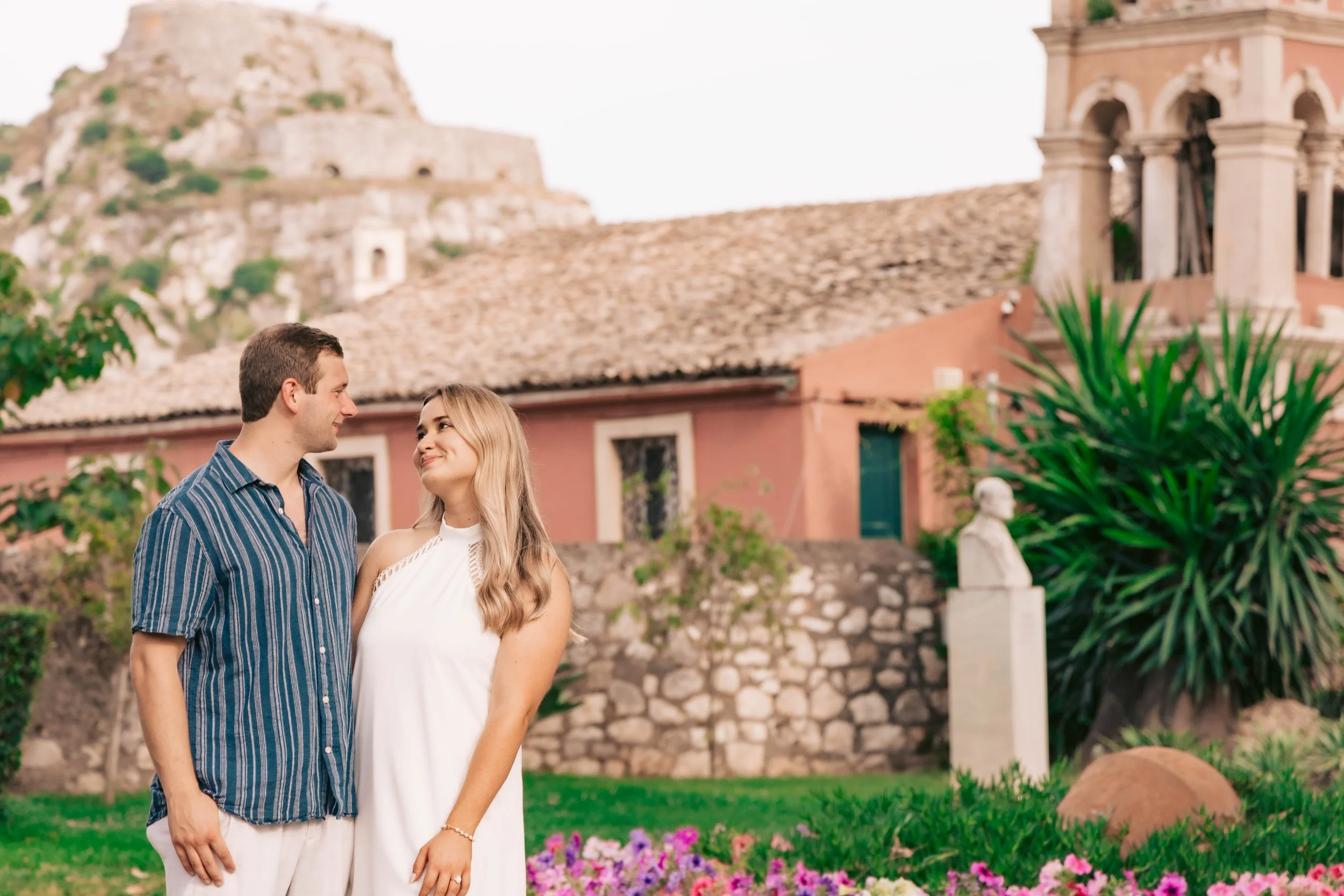 A young couple standing close together in a garden, looking at each other and smiling, with an old building and greenery in the background.