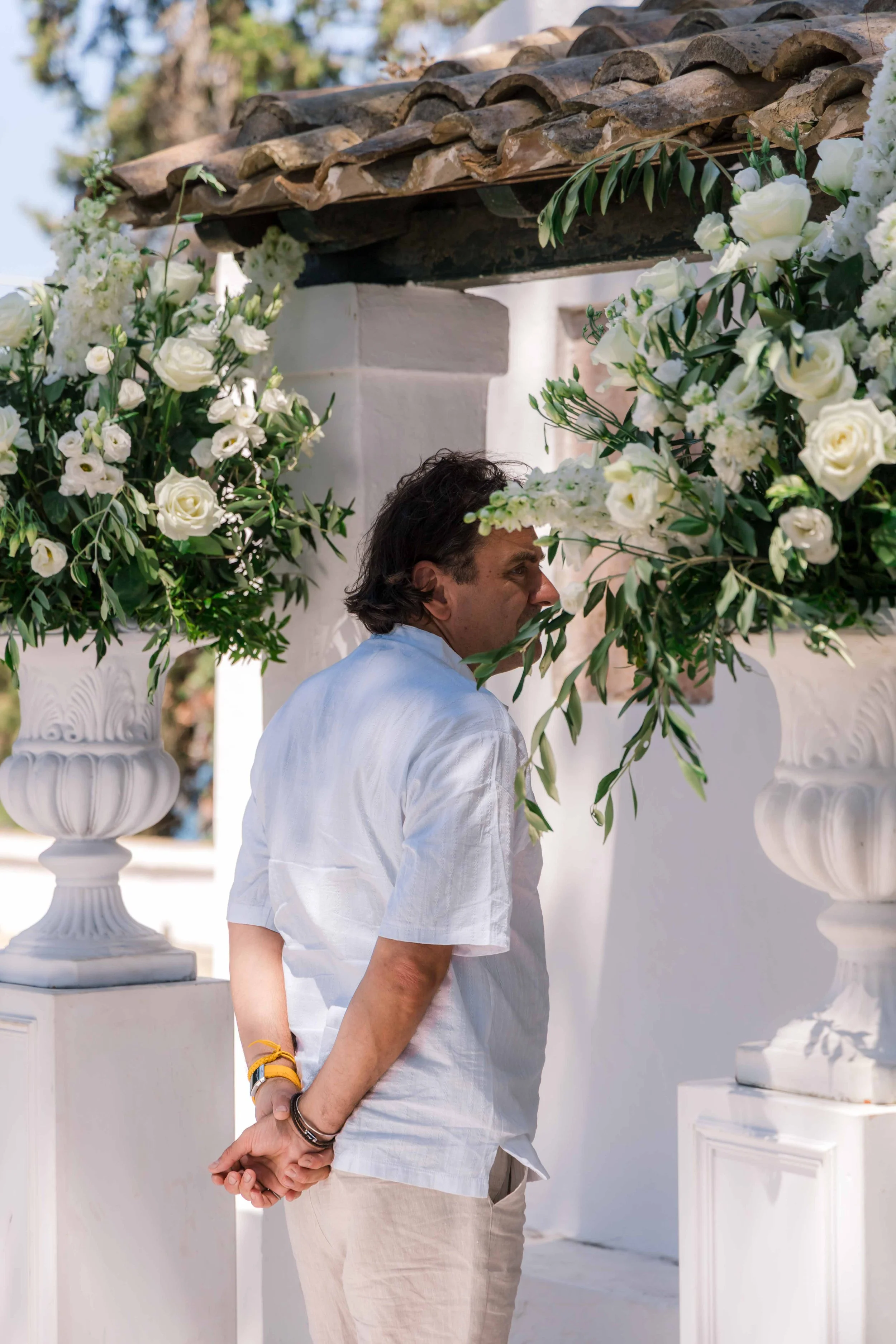 A man in a white shirt and beige pants standing with his hands behind his back near large white flower arrangements in vases.