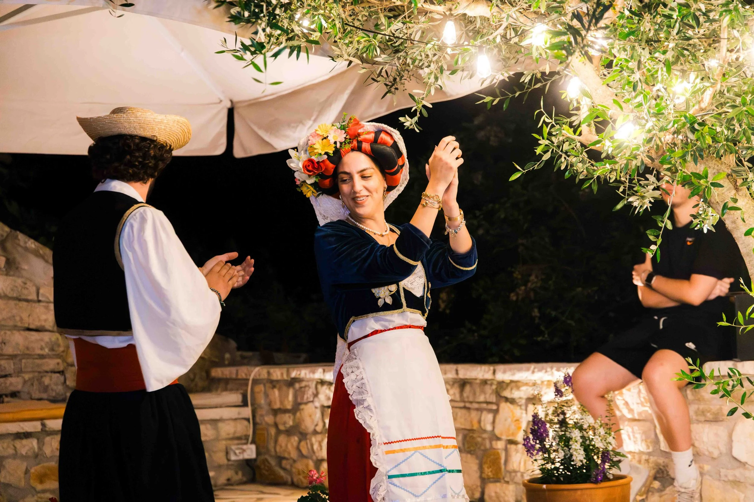 People in traditional clothing dancing outdoors at night, with string lights and a tree in the background.
