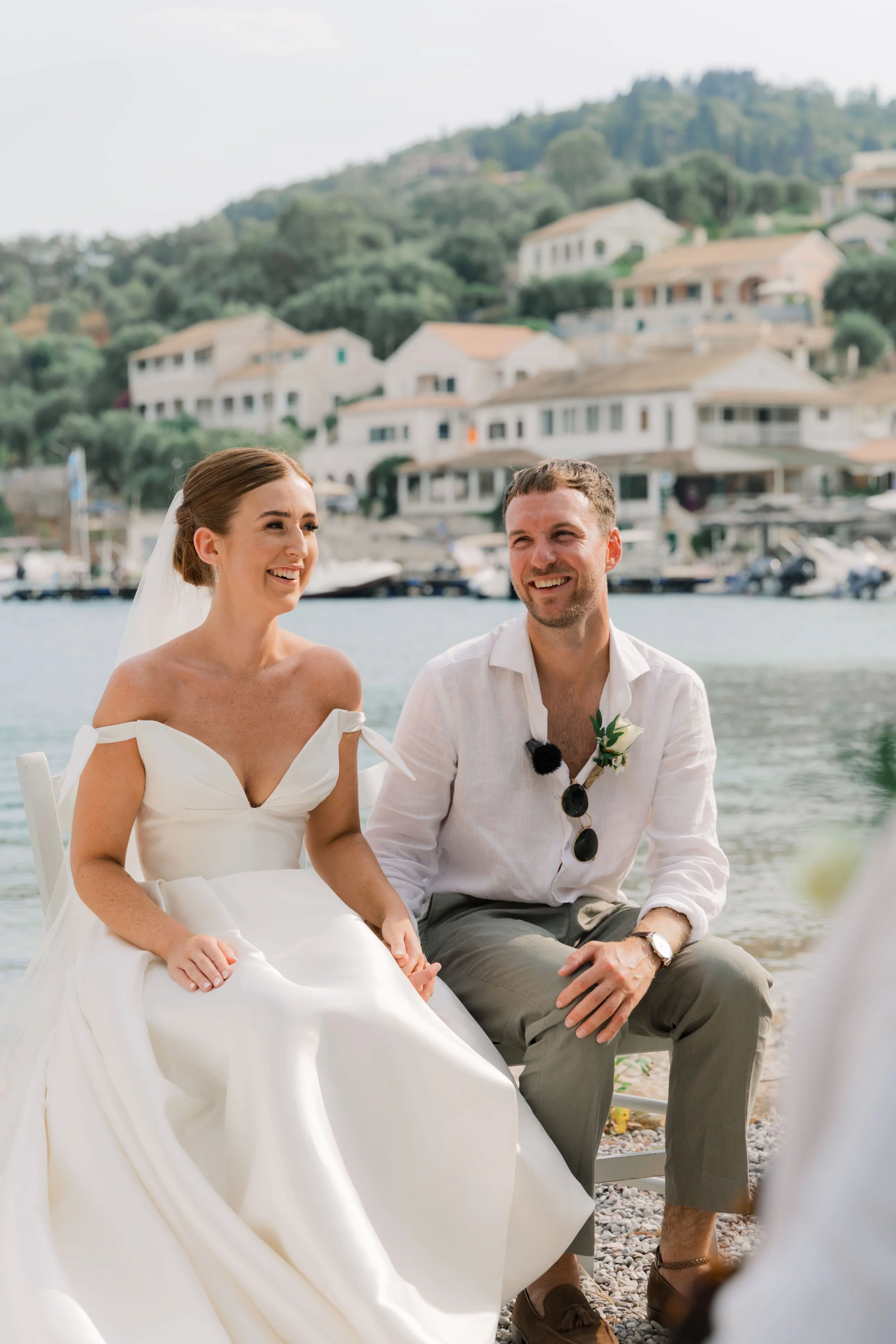 A woman in a white wedding dress and a man in white shirt and gray pants sitting near water, smiling, with hillside houses and boats in the background.