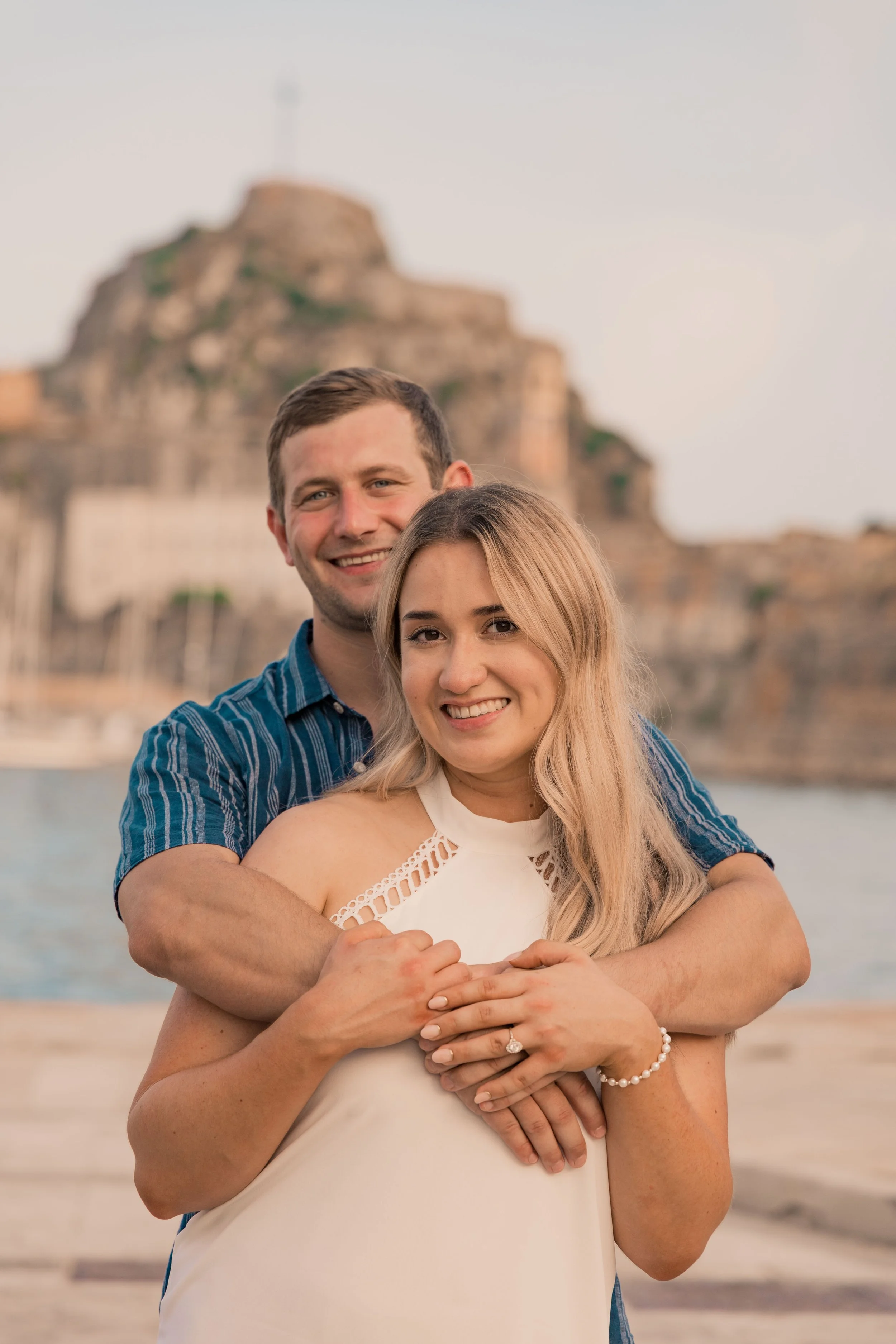 A smiling couple embracing, with a historic fort and waterfront in the background, during sunset.