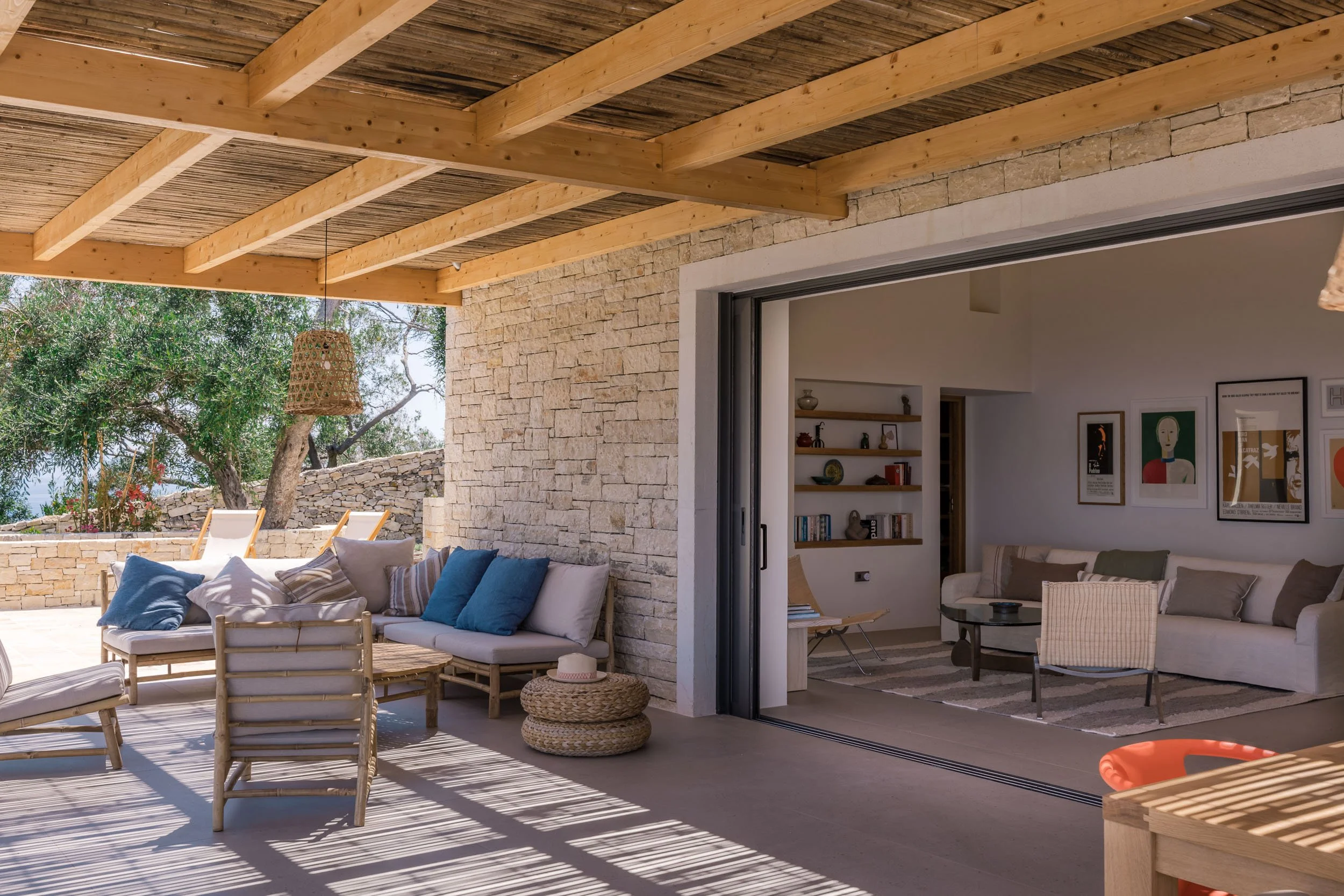 Indoor-outdoor living space with stone walls, wooden ceiling, and outdoor furniture including chairs, cushions, and woven ottomans, leading into a cozy living room with shelves, artwork, and a couch.