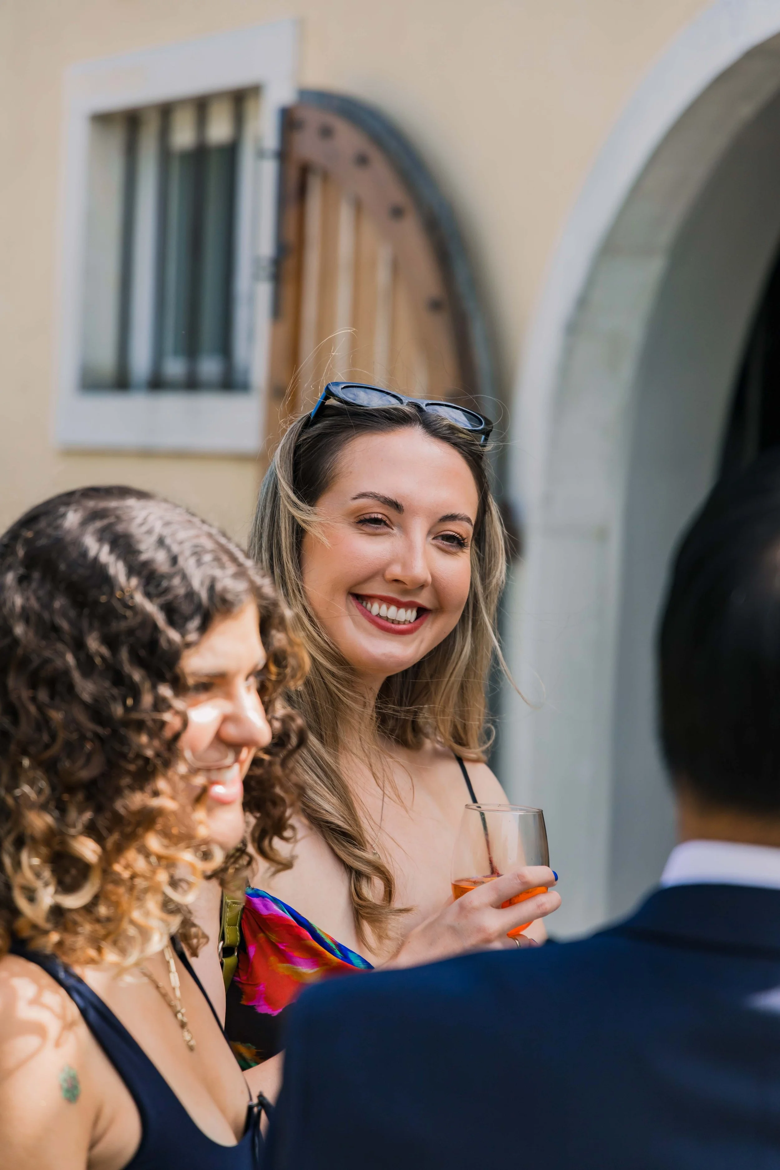 Two women smiling, one holding a glass of rosé wine, at an outdoor gathering with a European-style doorway and window in the background.