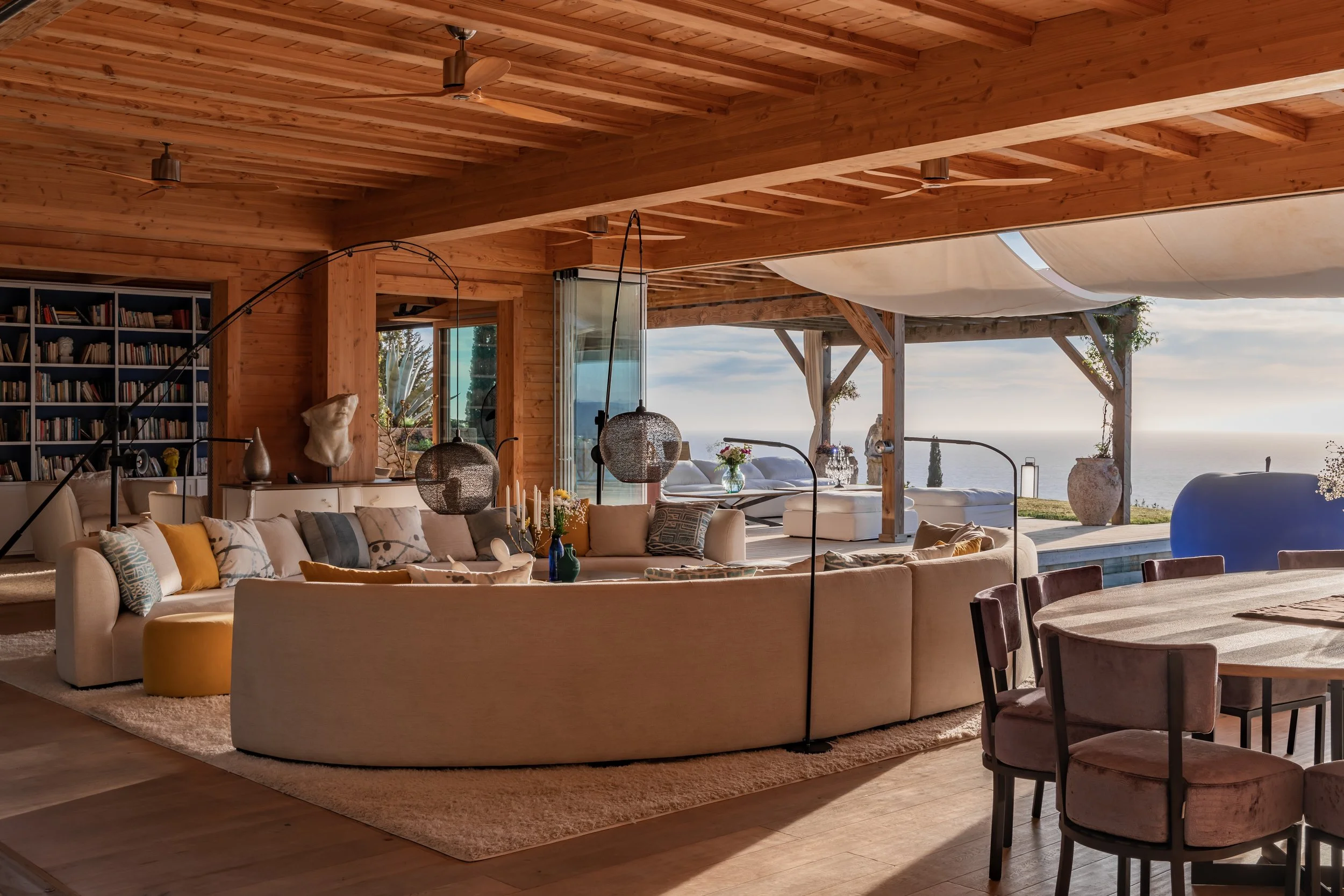 Interior of a modern living room with a circular sofa, table, and chairs, overlooking a patio with sea view, decorated with vases, books, and art pieces, featuring wooden ceiling and open sliding doors.