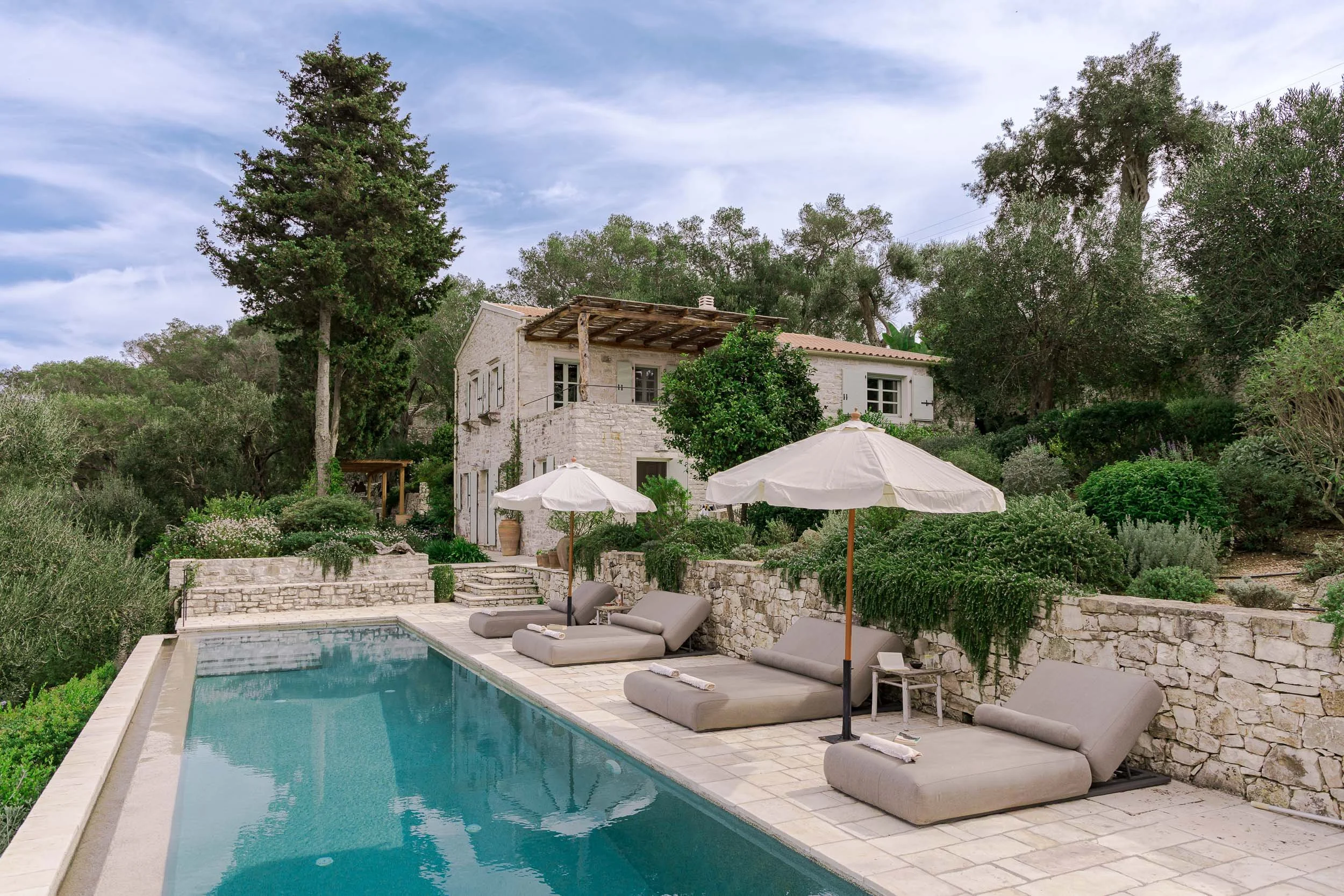 A luxurious outdoor pool area with three lounge chairs, and two white umbrellas, surrounded by stone walls and lush greenery, with a white house in the background.