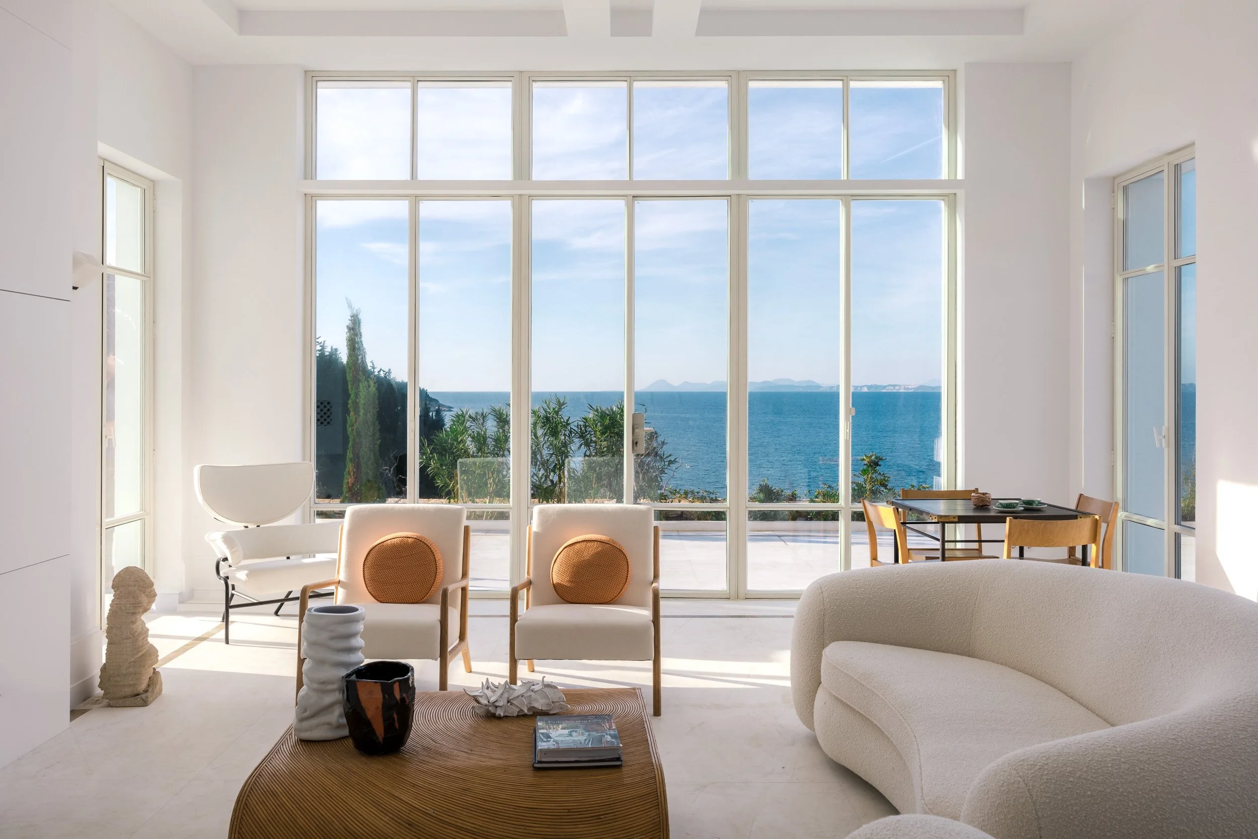Living room with large floor-to-ceiling windows overlooking the ocean, featuring white walls, contemporary furniture, and decorative items.